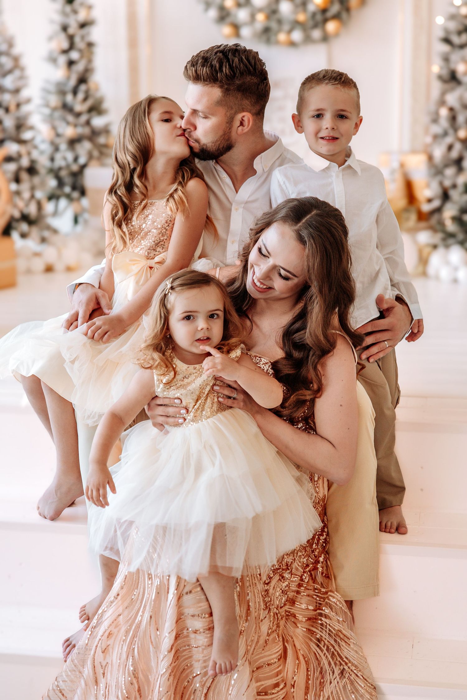 Family of five poses by Christmas trees, parents kissing daughter while son stands nearby, all dressed in elegant gold outfits.