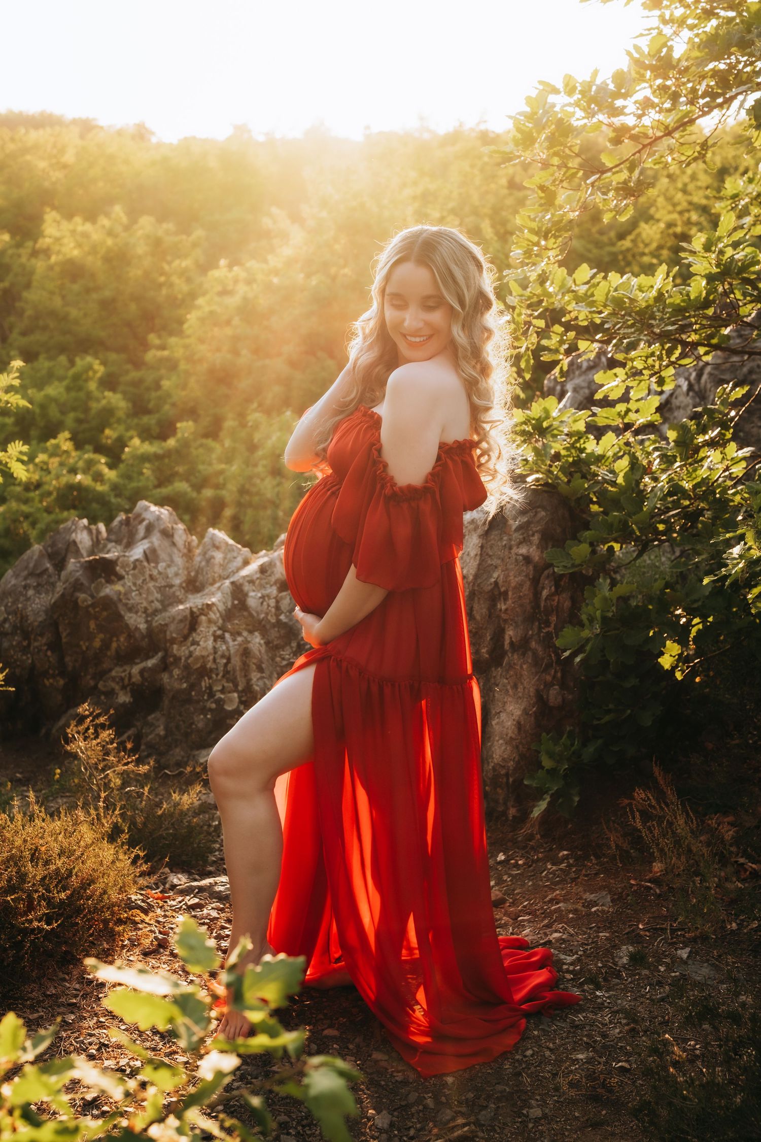 Happy pregnant woman in red maternity dress posing in lush green nature at sunset.