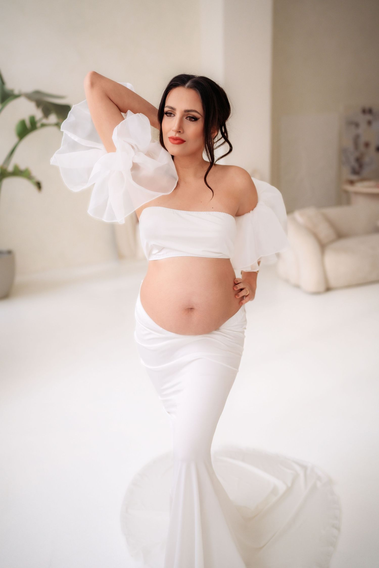 Pregnant woman in white off-shoulder top and mermaid skirt posing in bright maternity studio.
