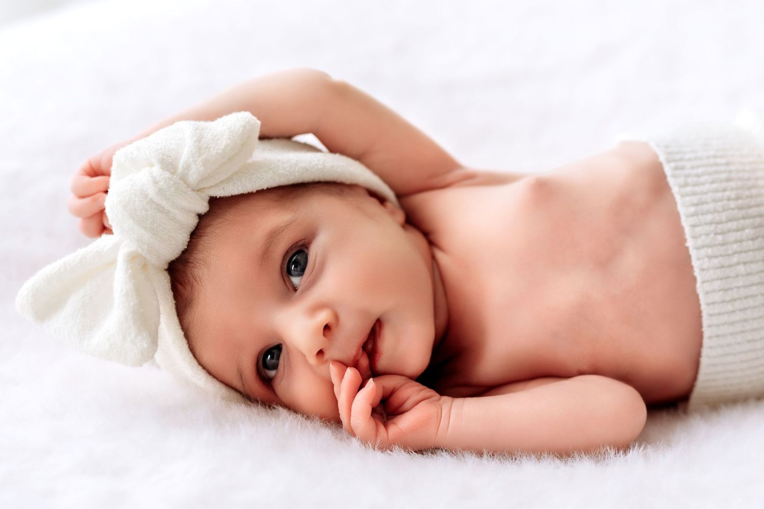 Adorable newborn baby with white bow headband lying on soft white blanket.
