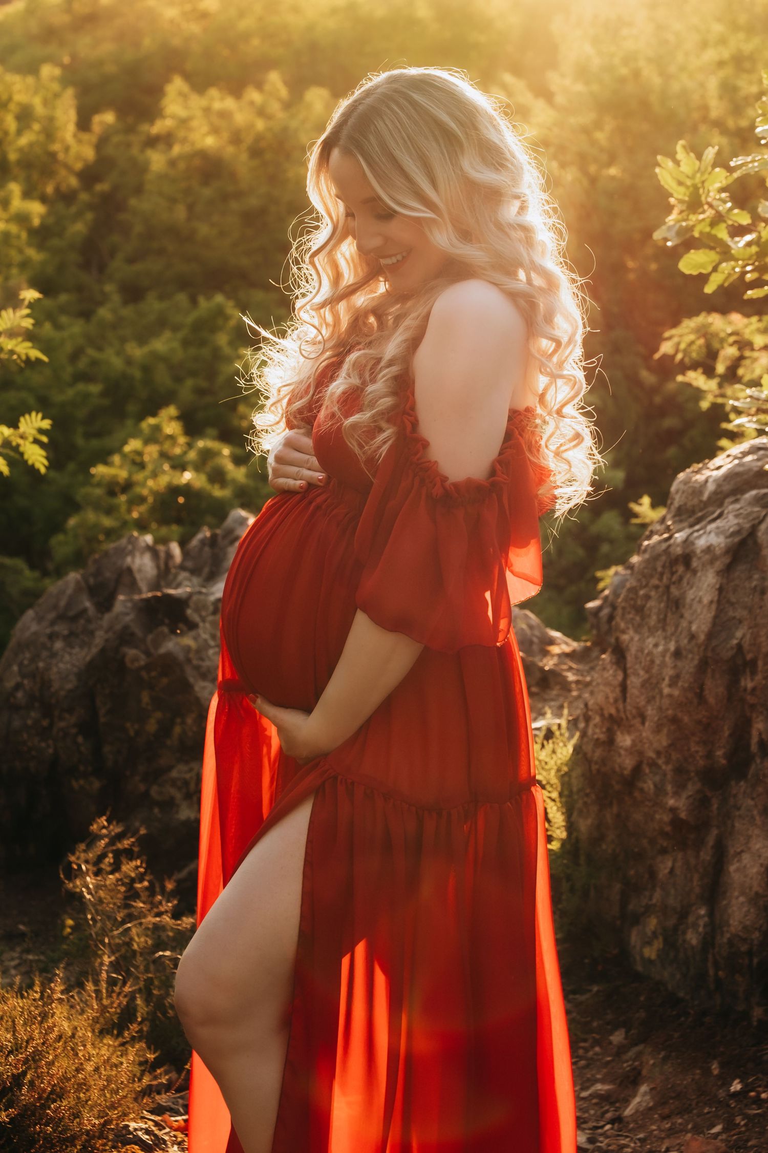 Smiling pregnant blonde woman in flowing red dress cradling bump in nature at sunset.
