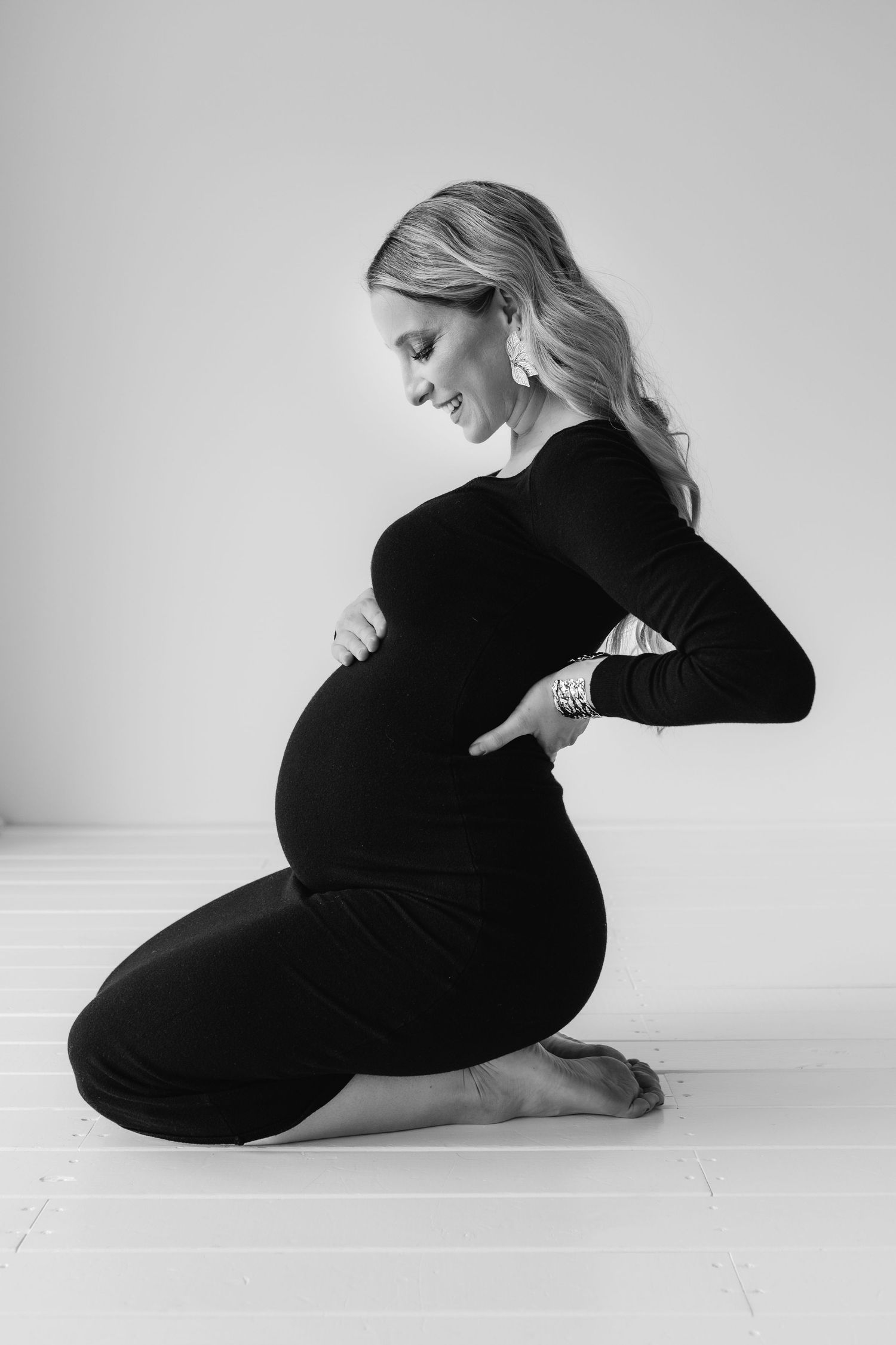 Black and white photo of pregnant woman in black dress kneeling on floor, lovingly holding her belly.
