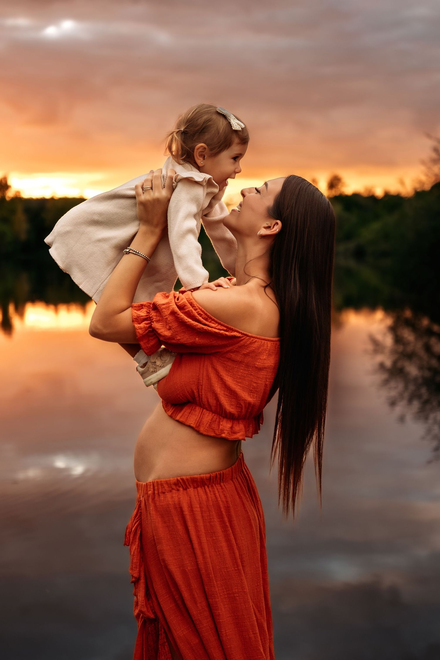 Pregnant brunette mother in red dress lifting smiling toddler girl against dramatic golden sunset lake backdrop.