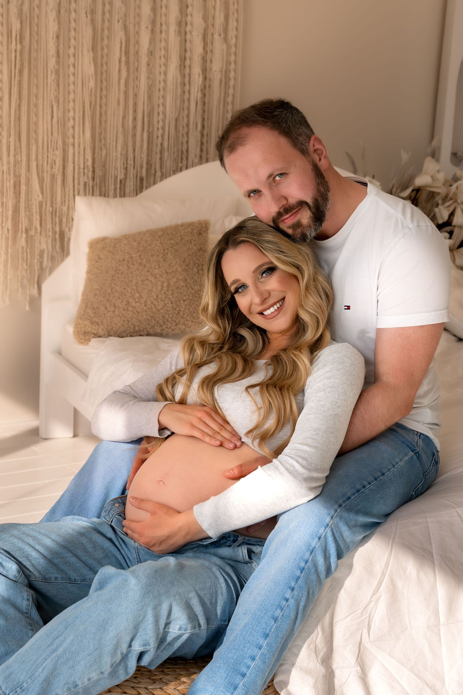 Happy couple sitting on bed, man embracing smiling pregnant woman while holding her baby bump.