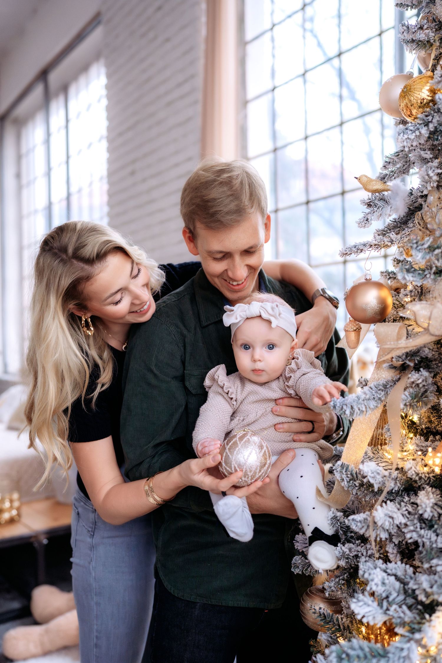 Young family with baby girl decorating Christmas tree with ornaments in bright living room.