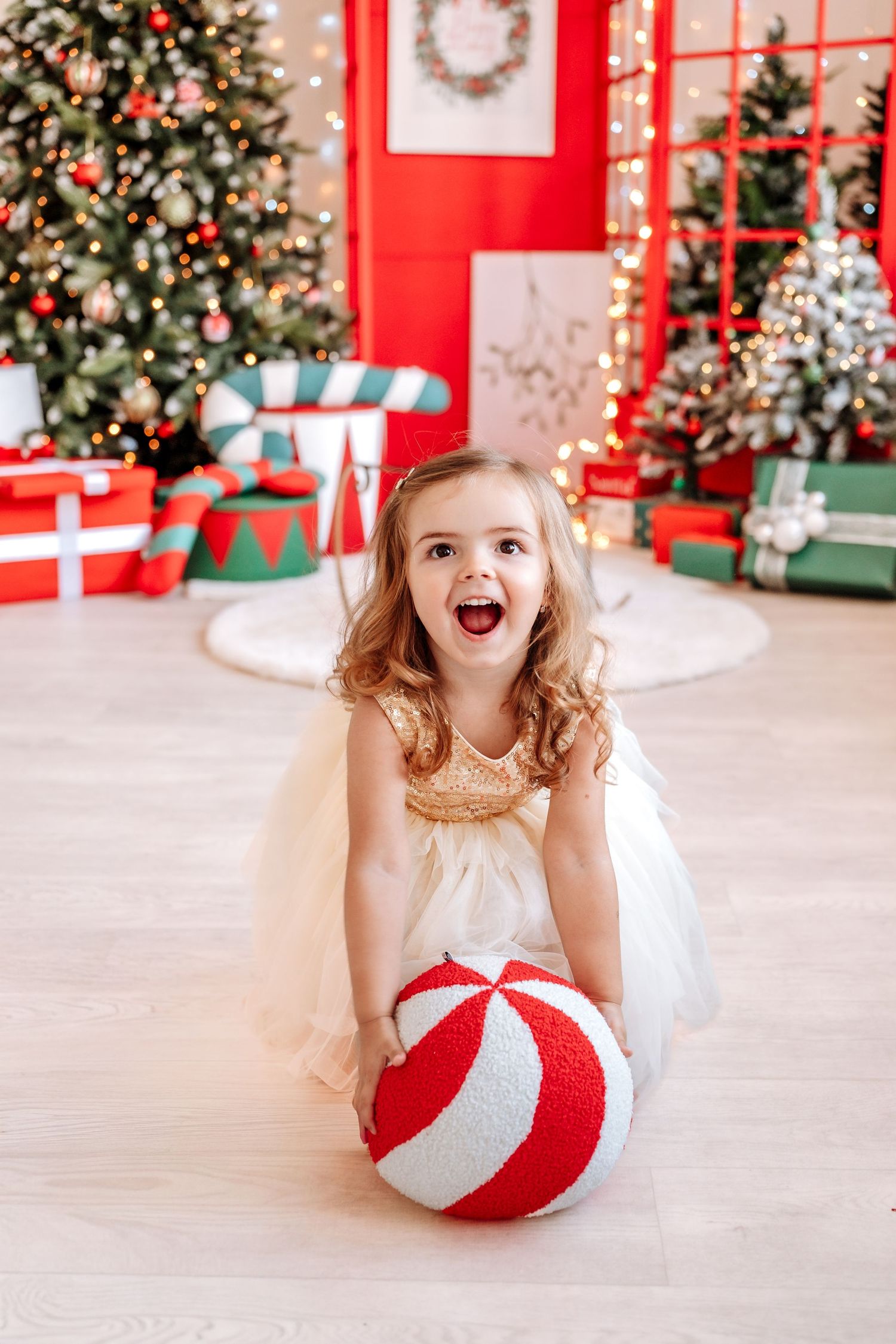 Excited little girl in white dress holding red and white striped ball near decorated Christmas tree with gifts.