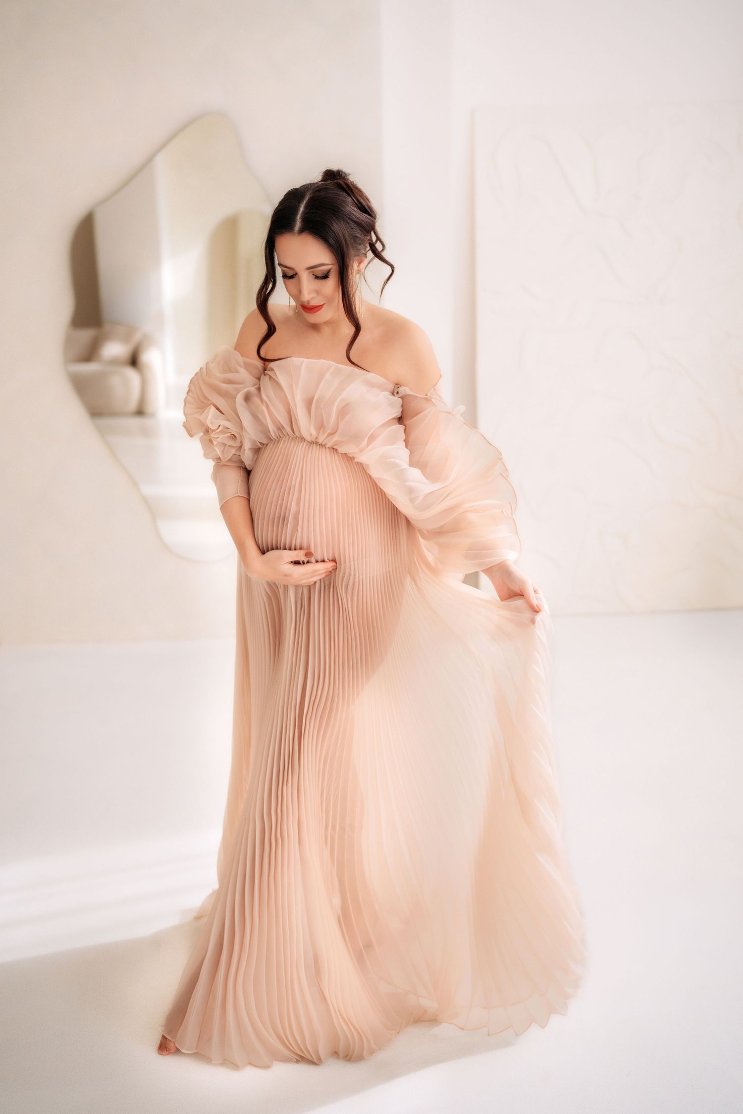Pregnant woman in flowing blush pleated off-shoulder gown looking down at her belly.