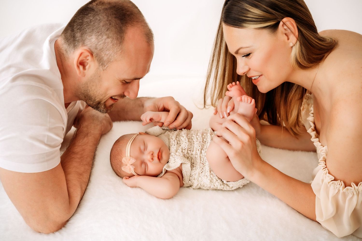 Parents lovingly admire their sleeping newborn baby girl in a soft white setting.