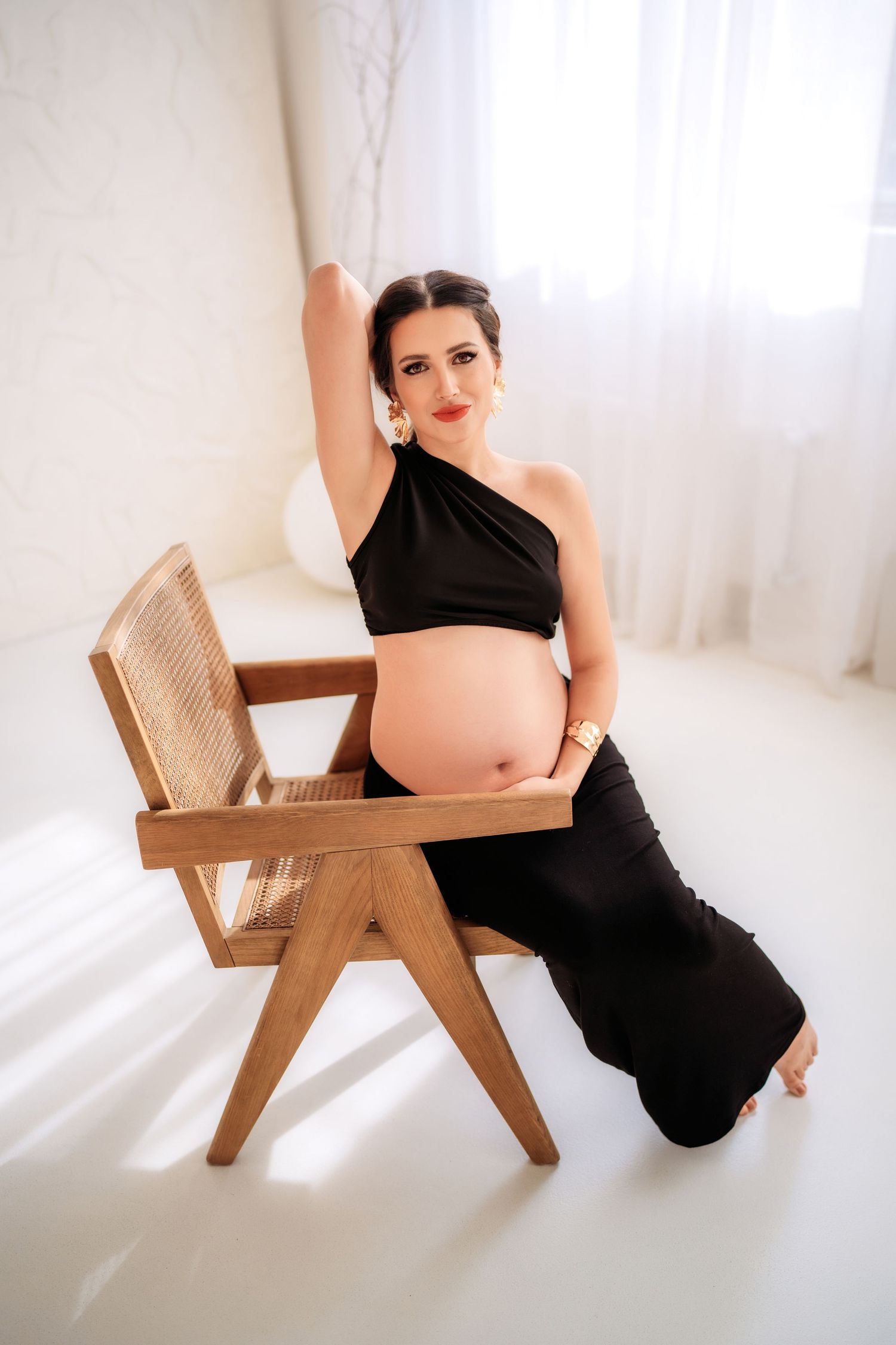Pregnant woman in black outfit sitting beside wooden chair on white floor in bright studio.