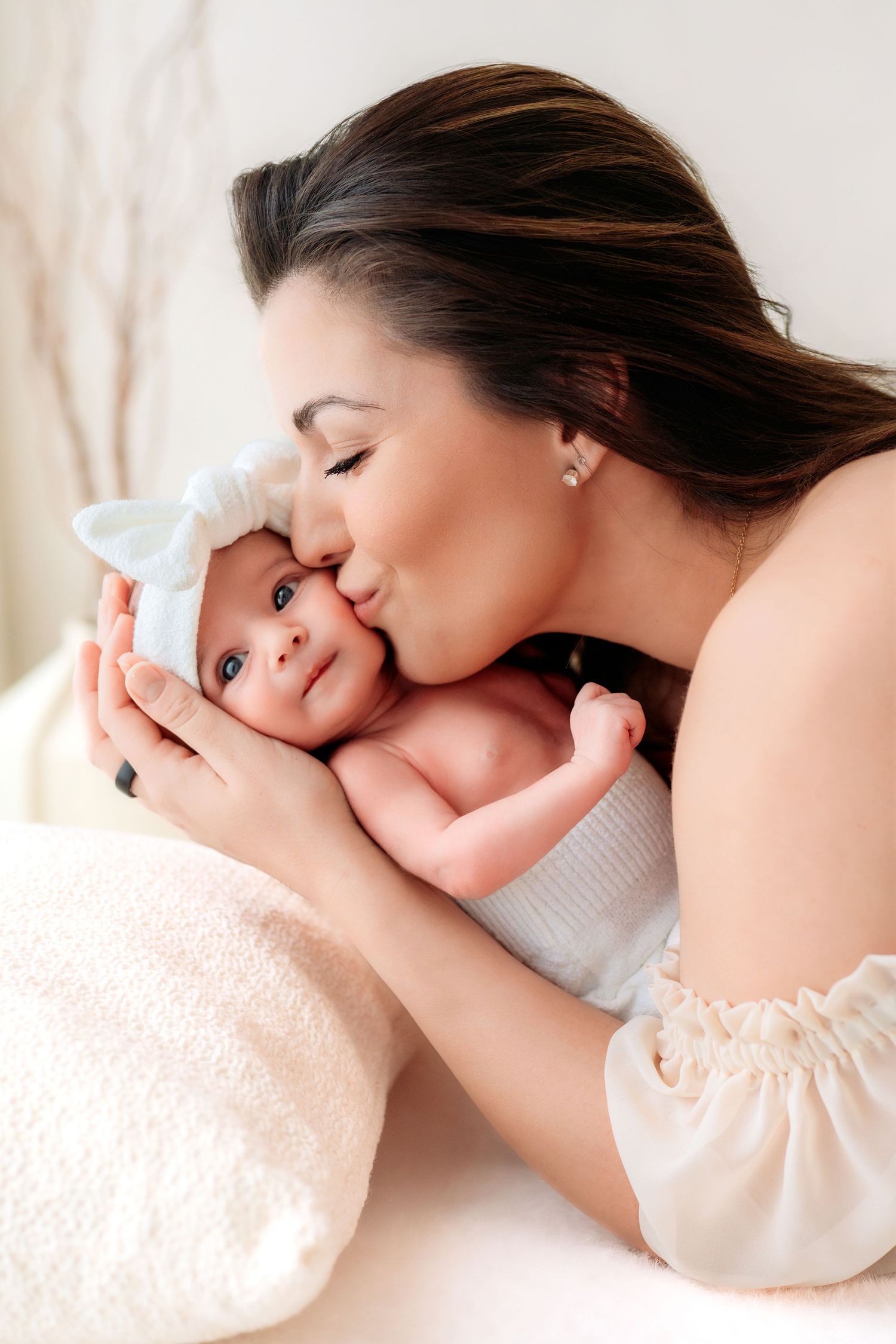Loving mother gently kissing her newborn baby wearing a white bow headband.