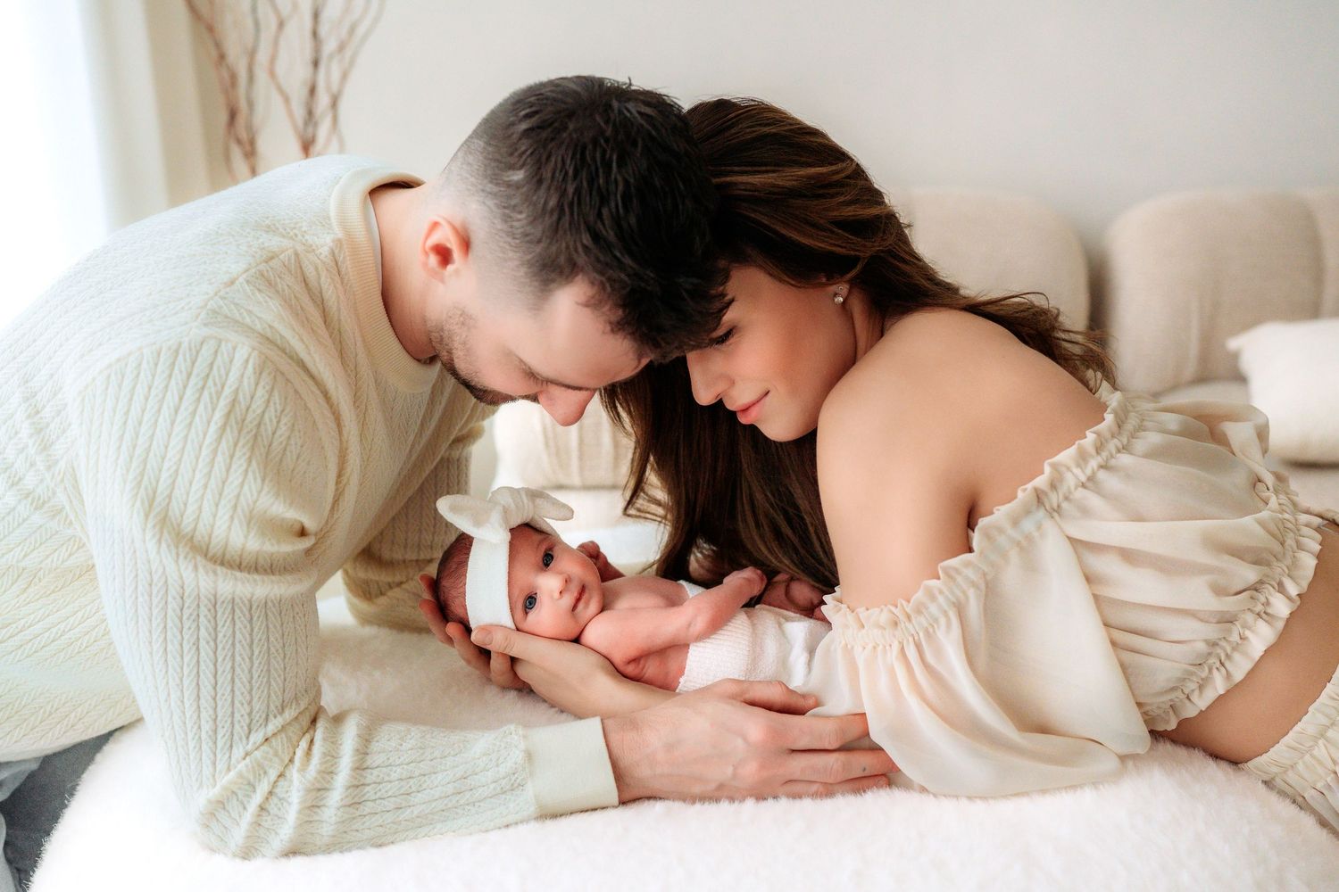 Parents leaning over white bed lovingly gazing at newborn baby girl with white bow headband.