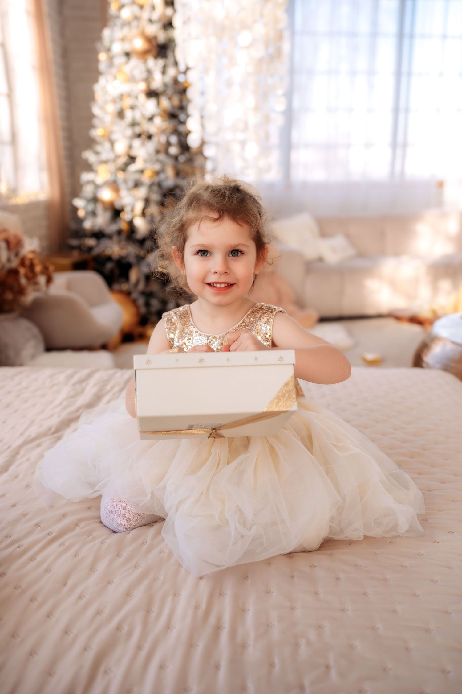 A smiling little girl in a gold sequin tulle dress sits on a bed holding a white gift box near a decorated Christmas tree.