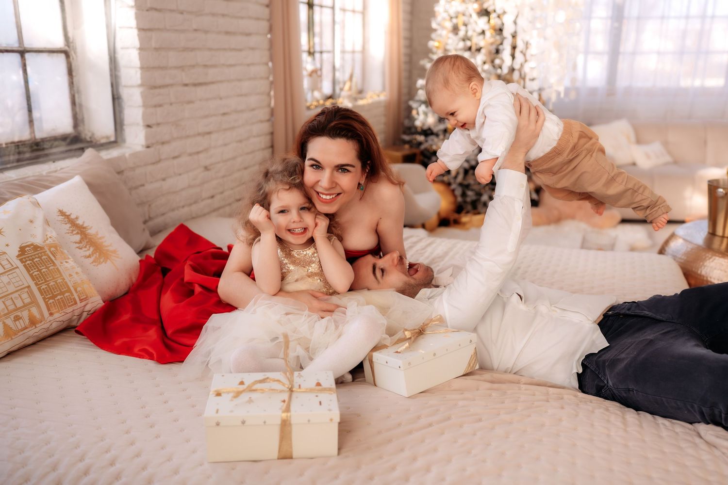 Mother smiling on bed with two young children surrounded by Christmas gifts and festive holiday decorations.