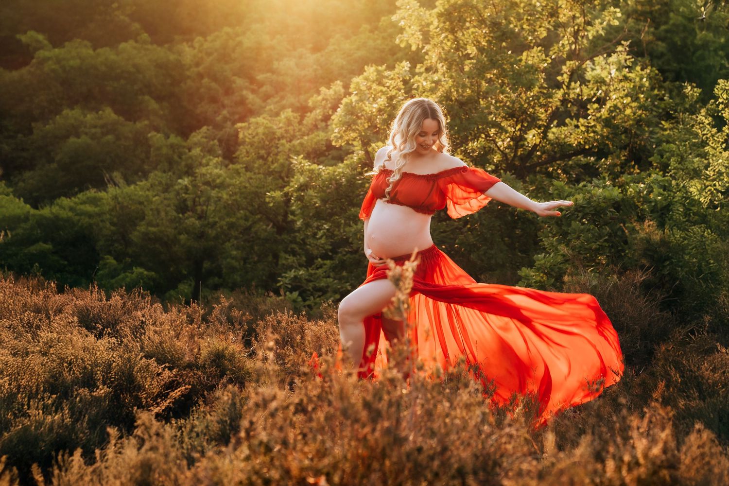 Pregnant woman in flowing red dress dancing in golden sunlight among wildflowers with lush green trees behind her.