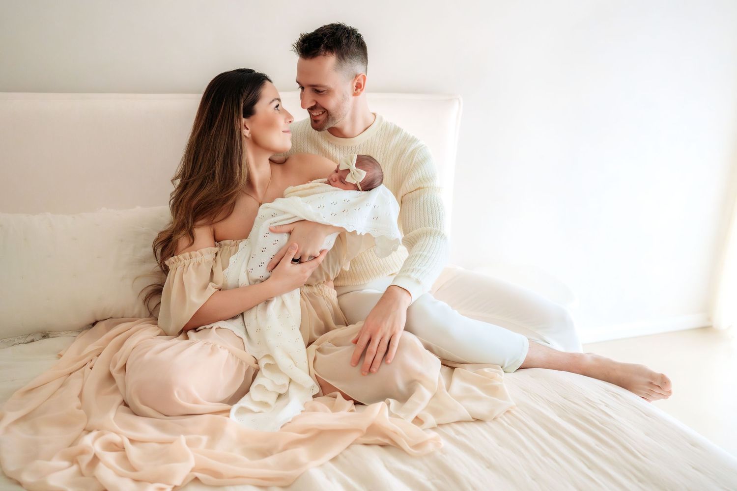 Smiling couple sitting on bed in neutral tones lovingly looking at their newborn baby together.