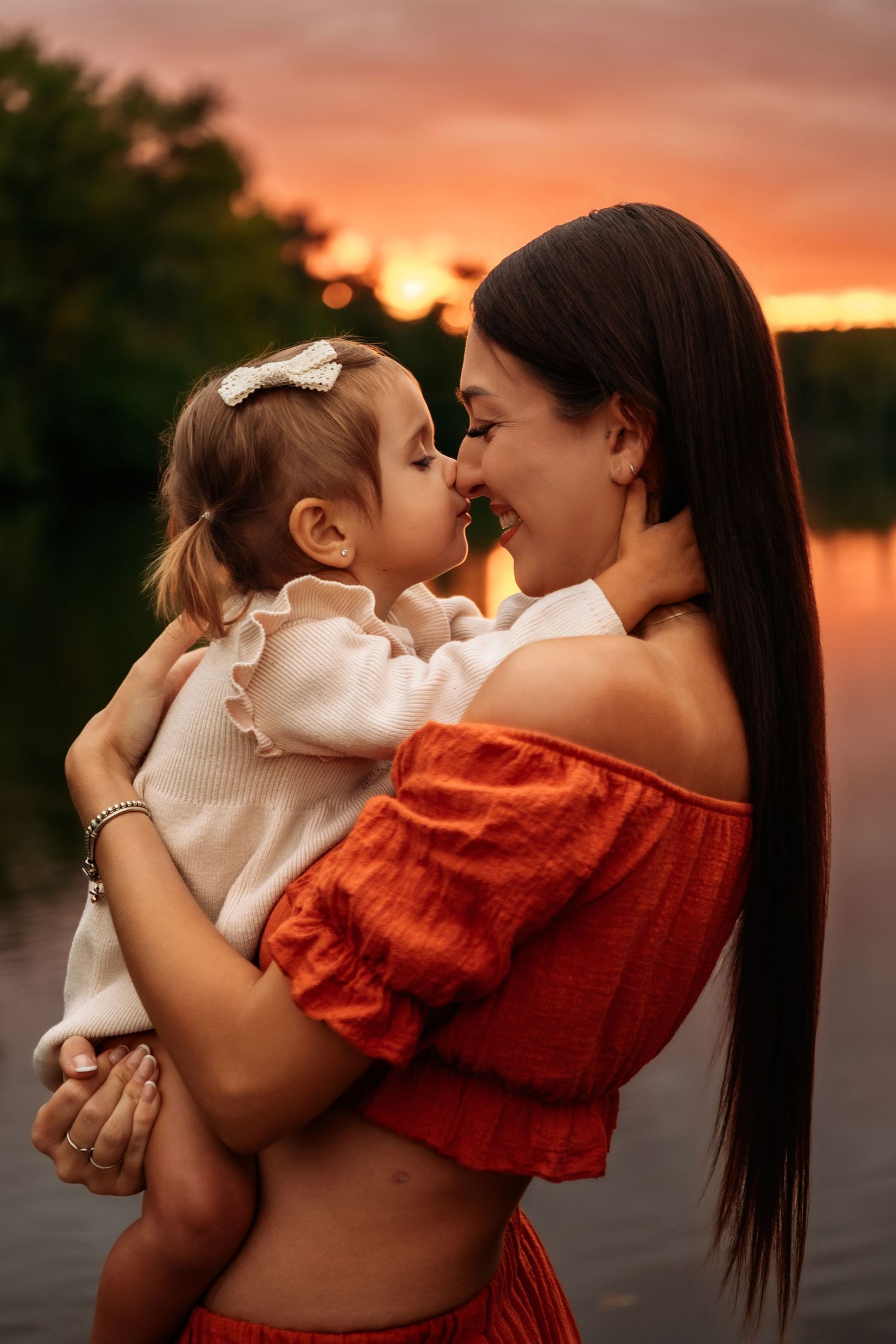 Mother and toddler daughter touch noses affectionately by lake at glowing red sunset.