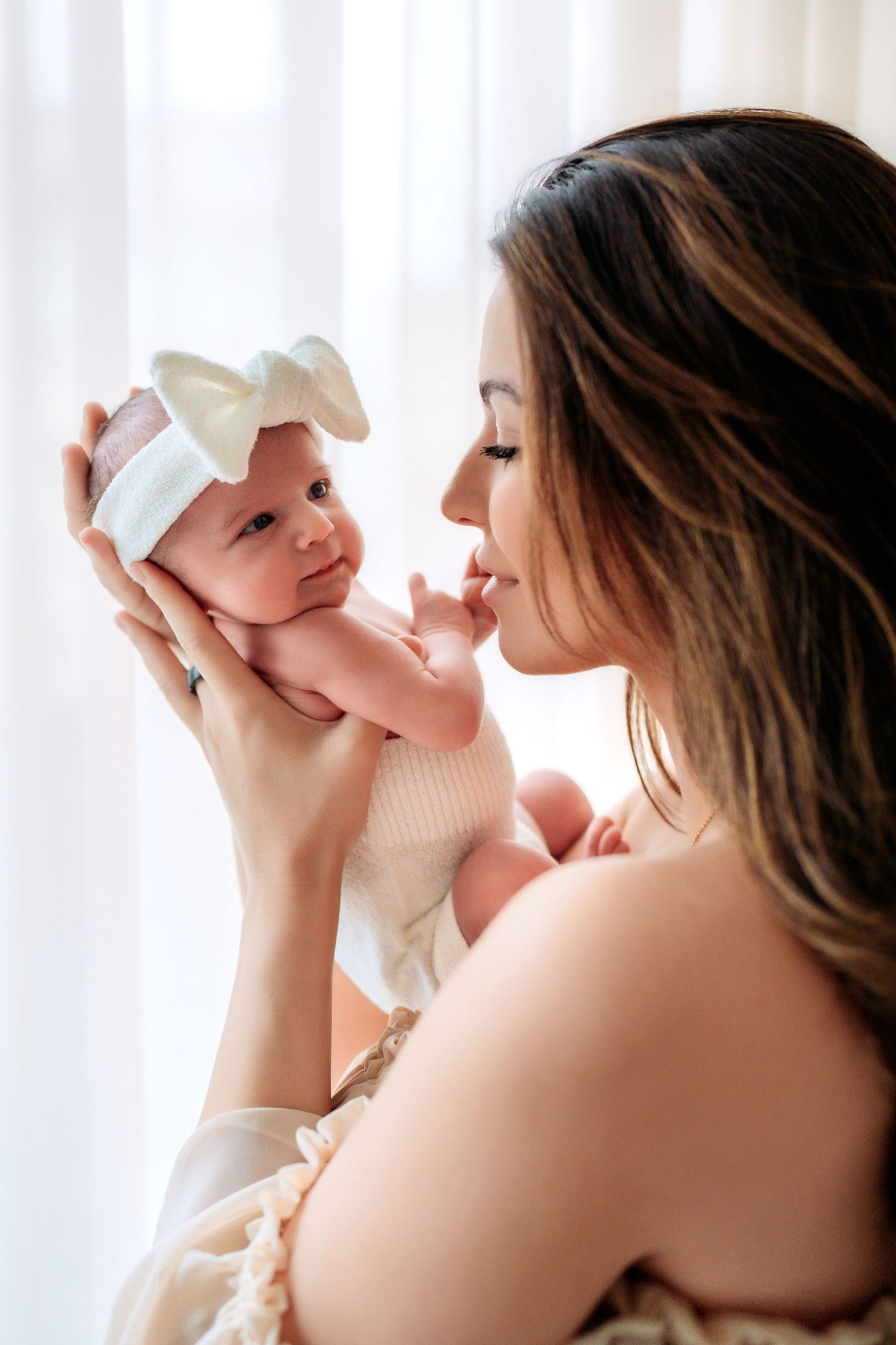 A brunette mother in a beige dress holds her newborn baby girl up close for a gentle kiss.
