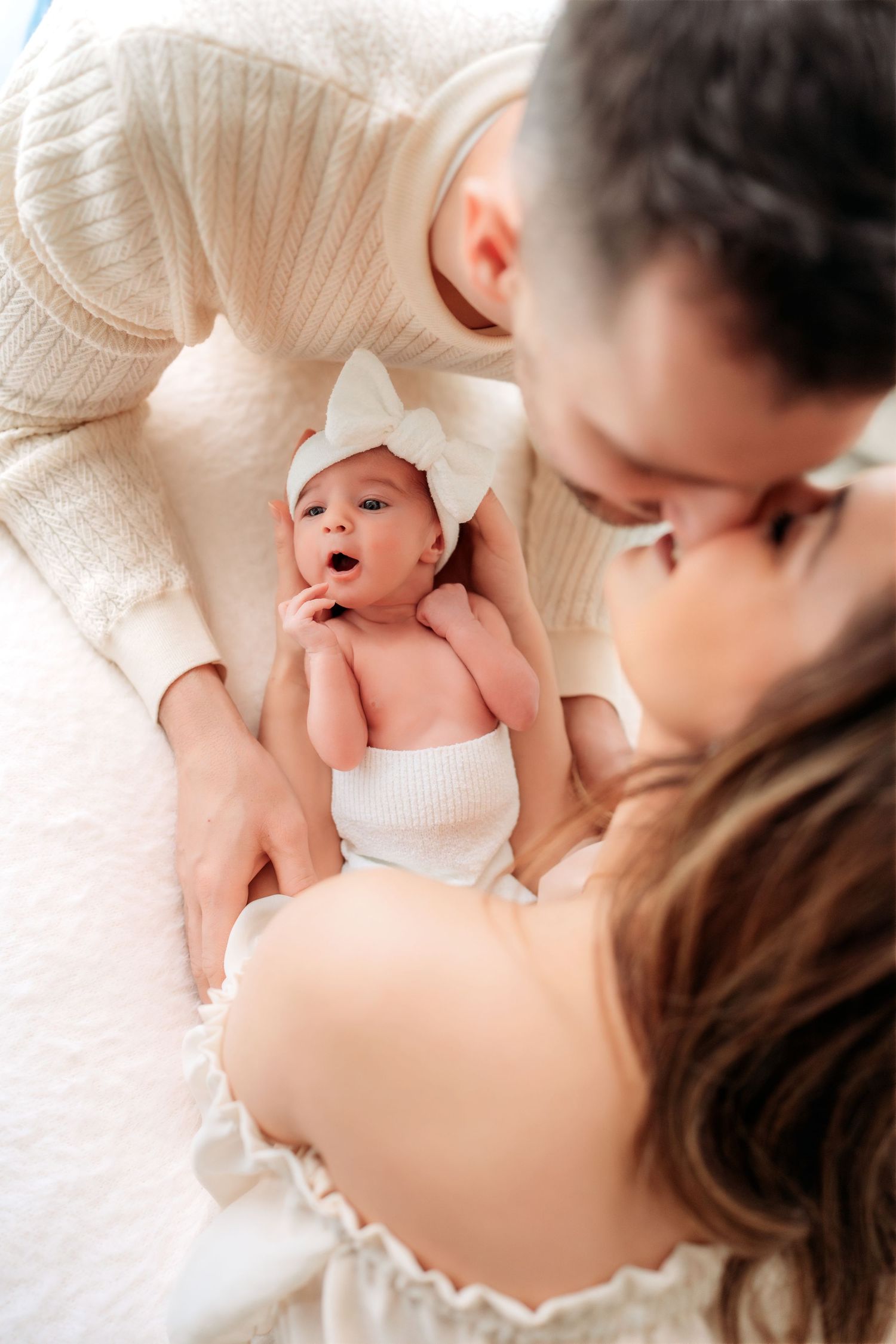 New parents in cream outfits lean in to kiss while their newborn baby girl lies between them.