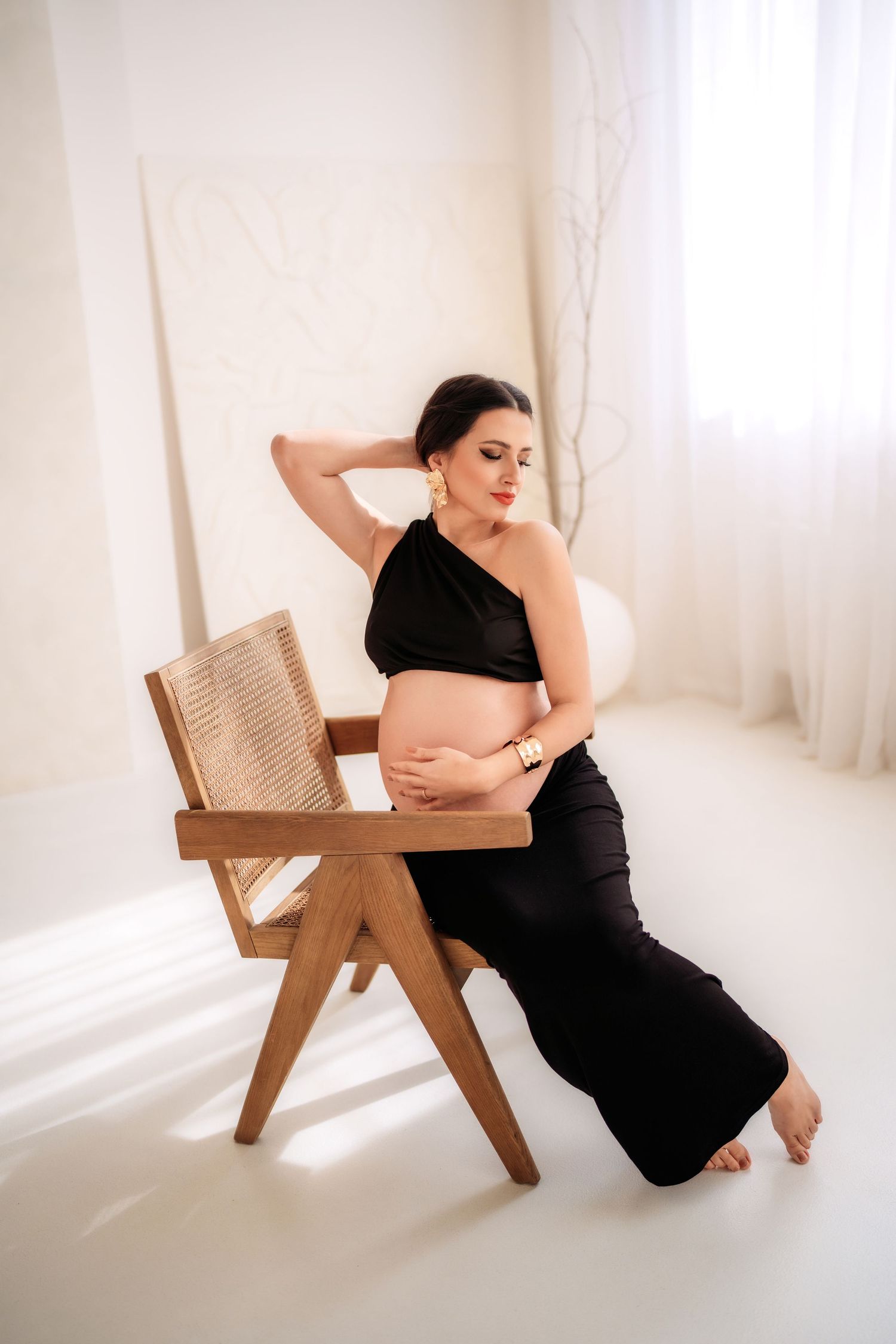 Pregnant woman in black crop top and pants sitting on wooden chair, cradling baby bump in bright white studio.