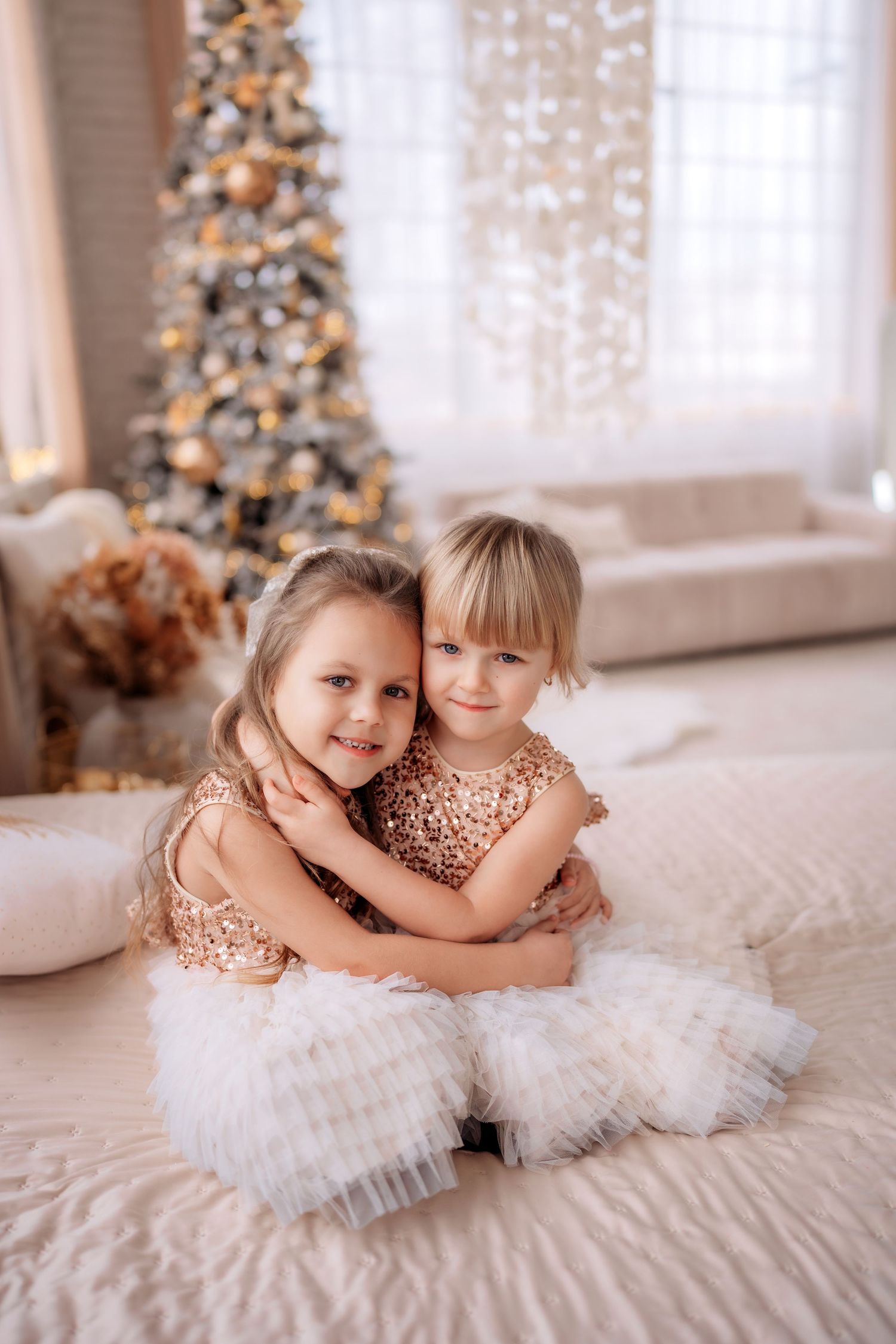 Two young girls in gold sequin dresses hugging on a bed in front of a decorated Christmas tree.
