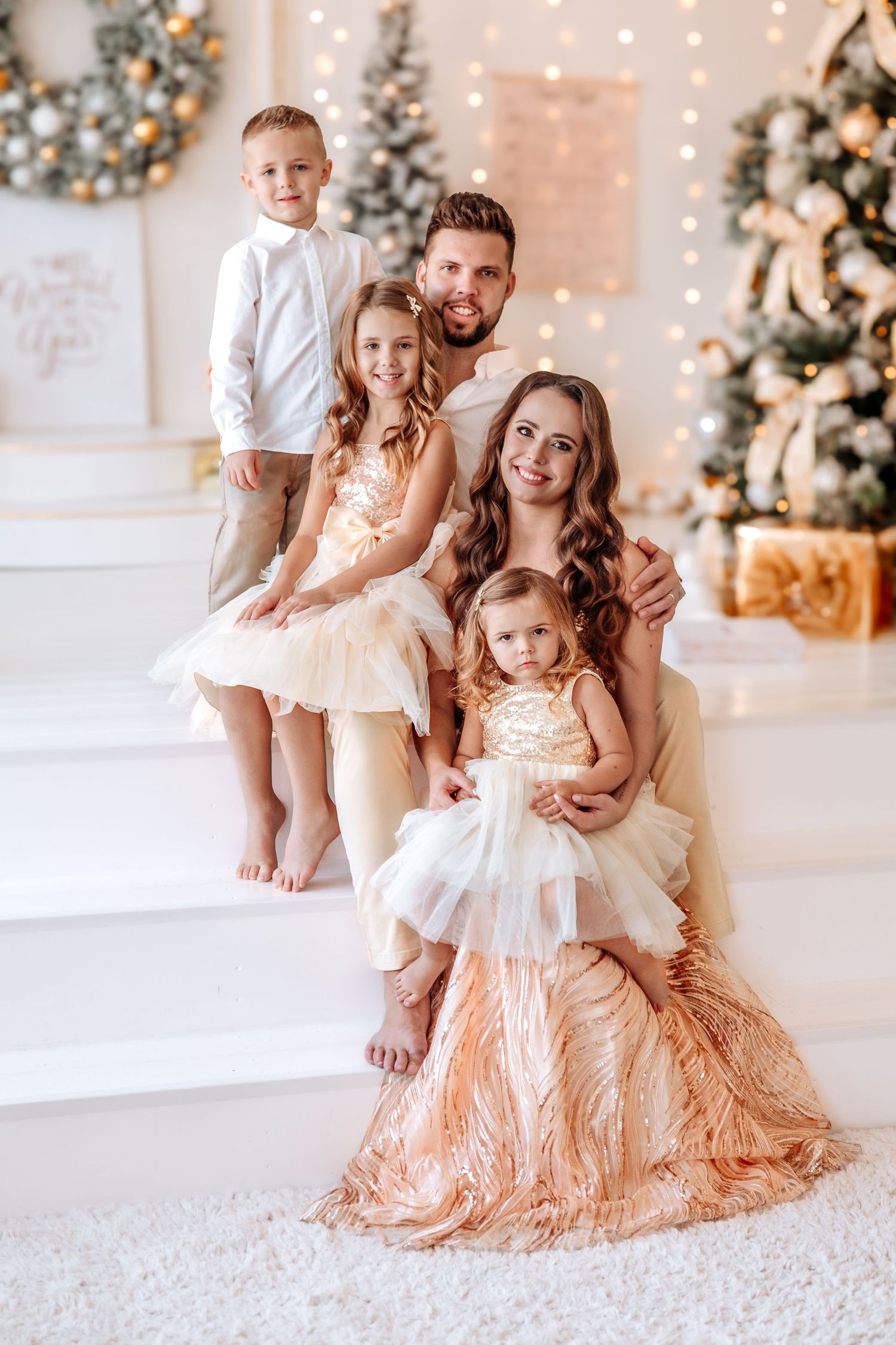 Happy family of four posing by decorated Christmas trees, wearing elegant gold and white holiday outfits.