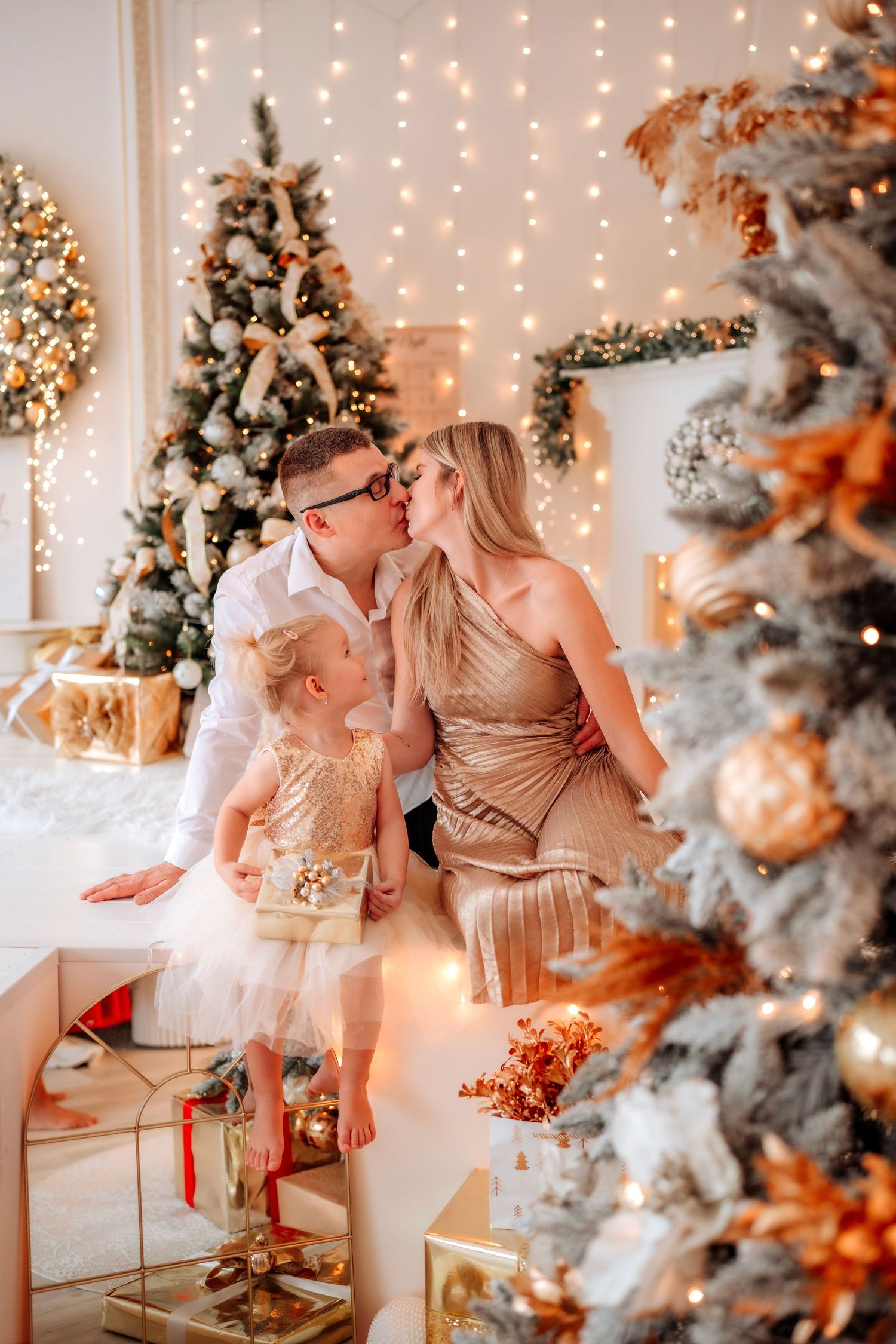 Family sharing a kiss by decorated Christmas trees with golden ornaments and warm fairy lights in a festive living room.