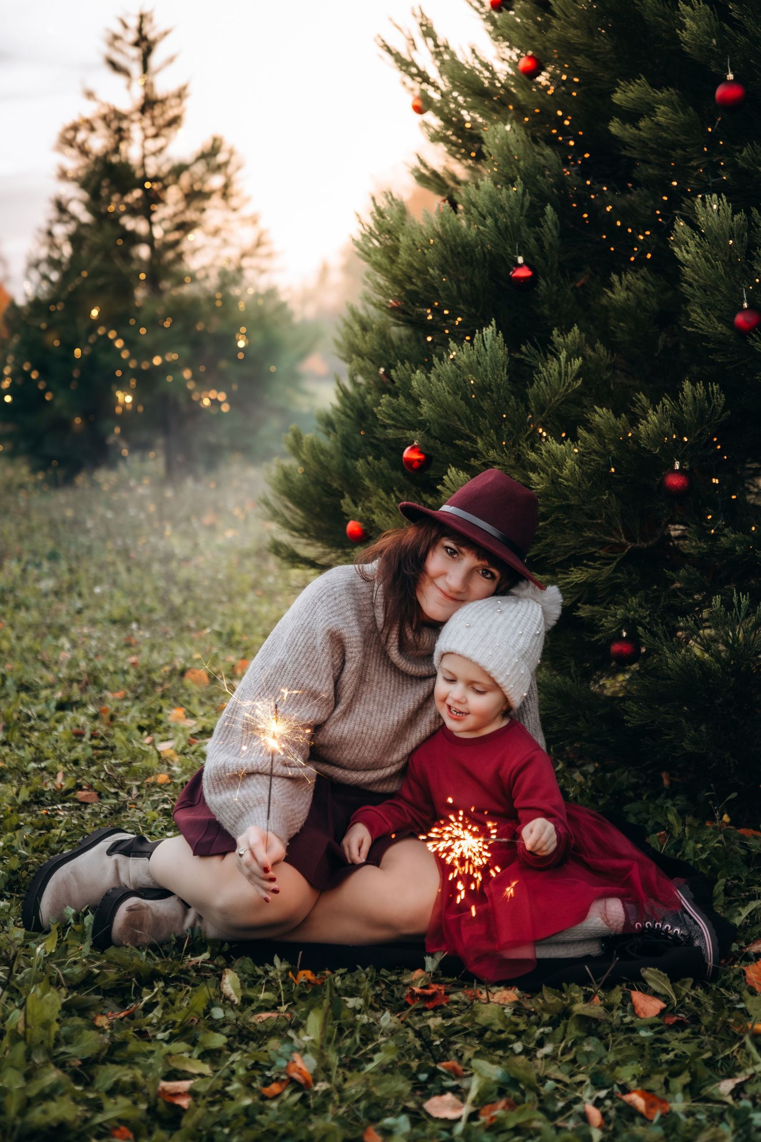 Mother and daughter sitting by decorated Christmas tree holding sparklers outdoors at sunset.