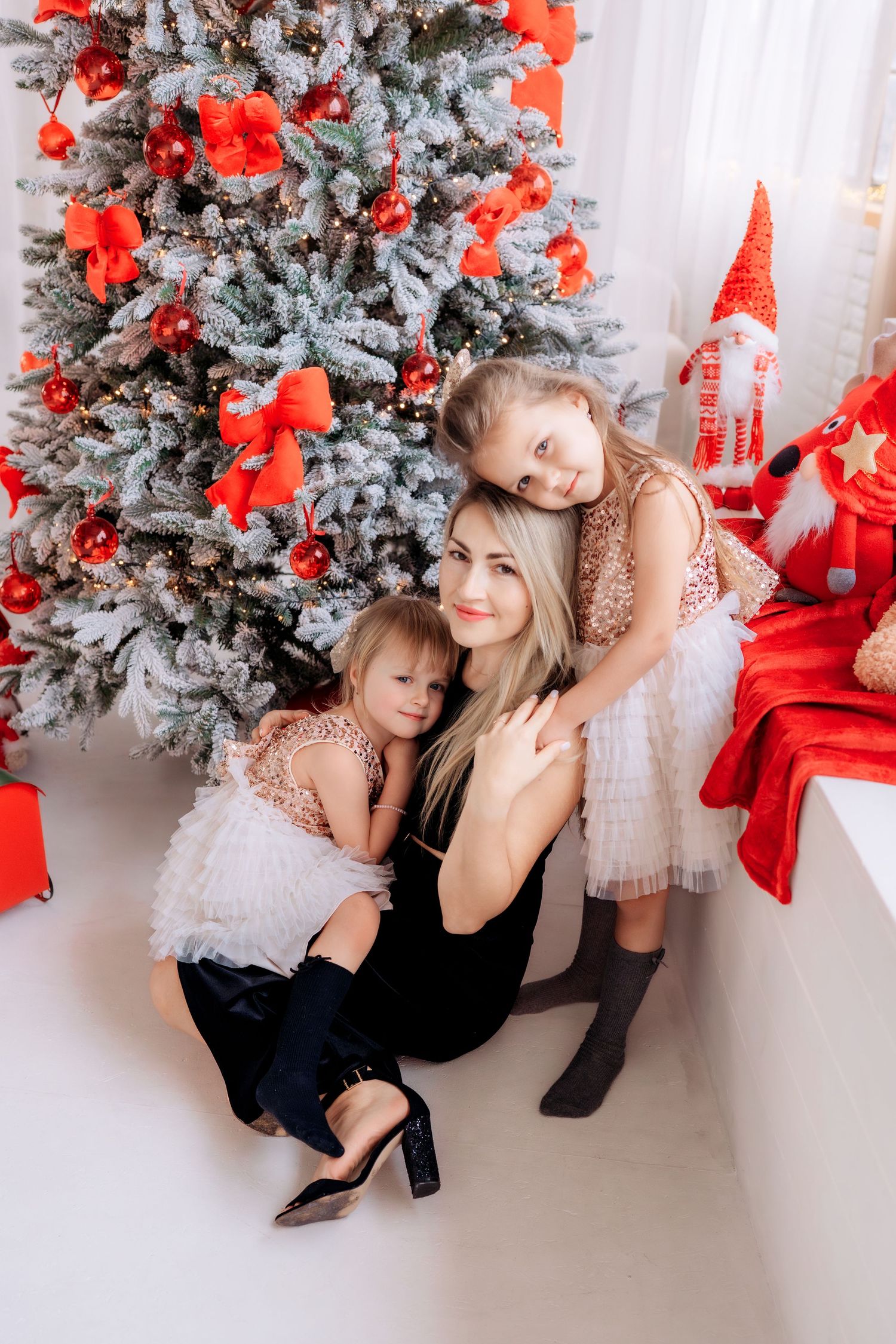 Mother and two daughters in festive dresses hugging by a decorated Christmas tree with red ornaments and ribbons.