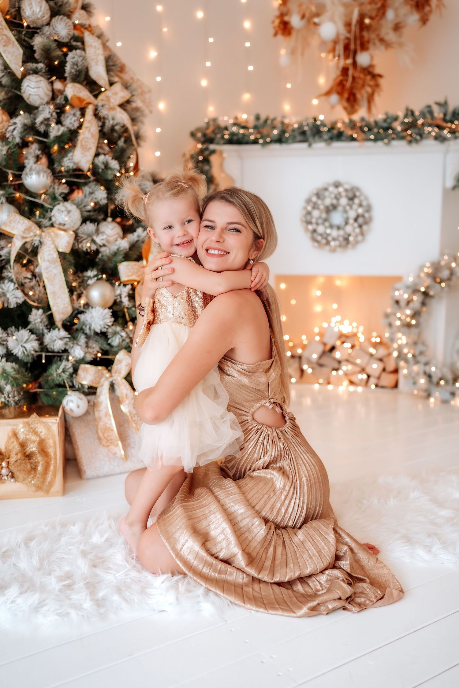 Mother and daughter hugging by a decorated Christmas tree with golden ornaments and warm glowing lights.