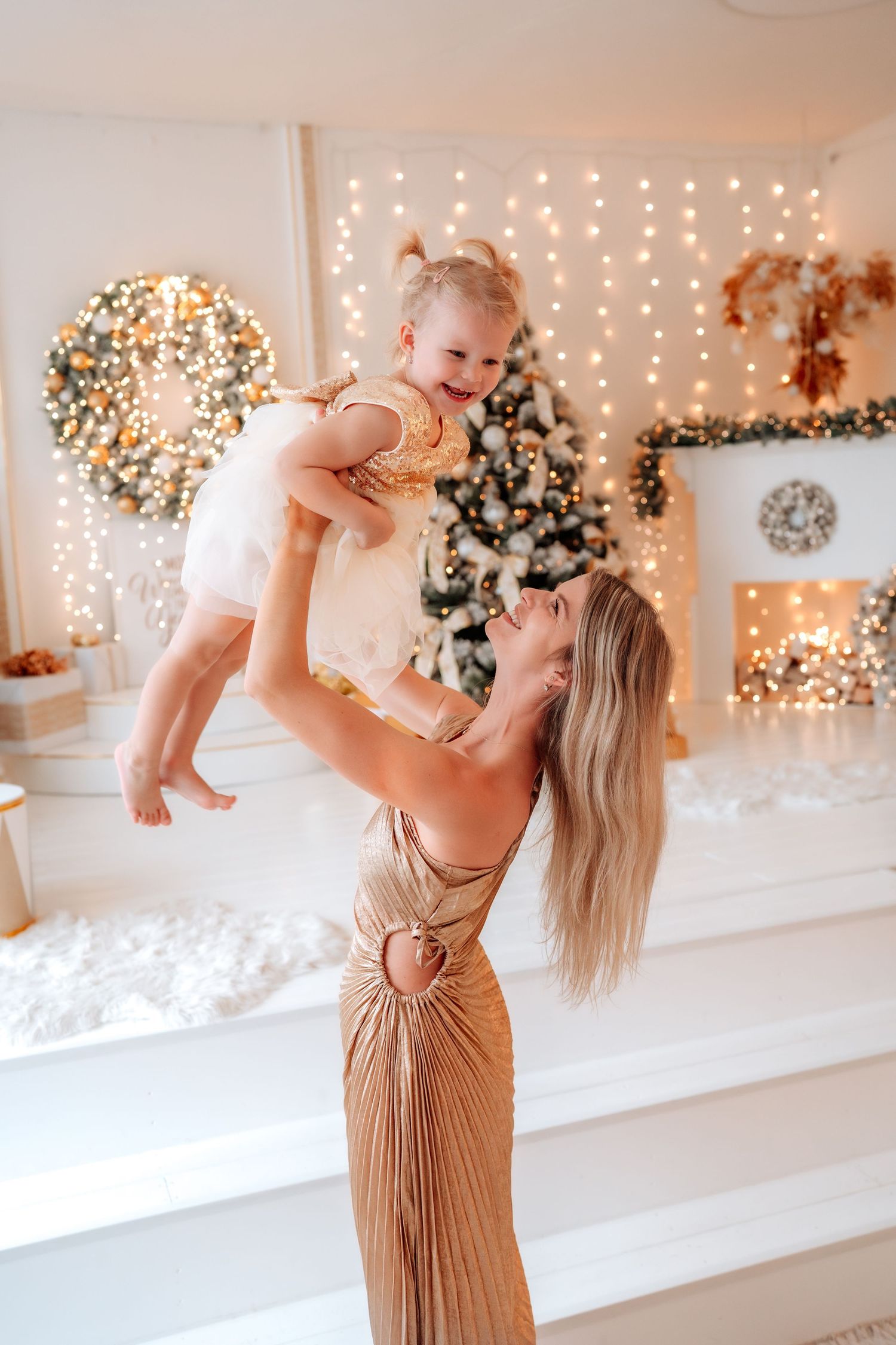 A joyful mother in a gold gown lifts her laughing baby girl near a decorated Christmas tree with warm glowing lights.