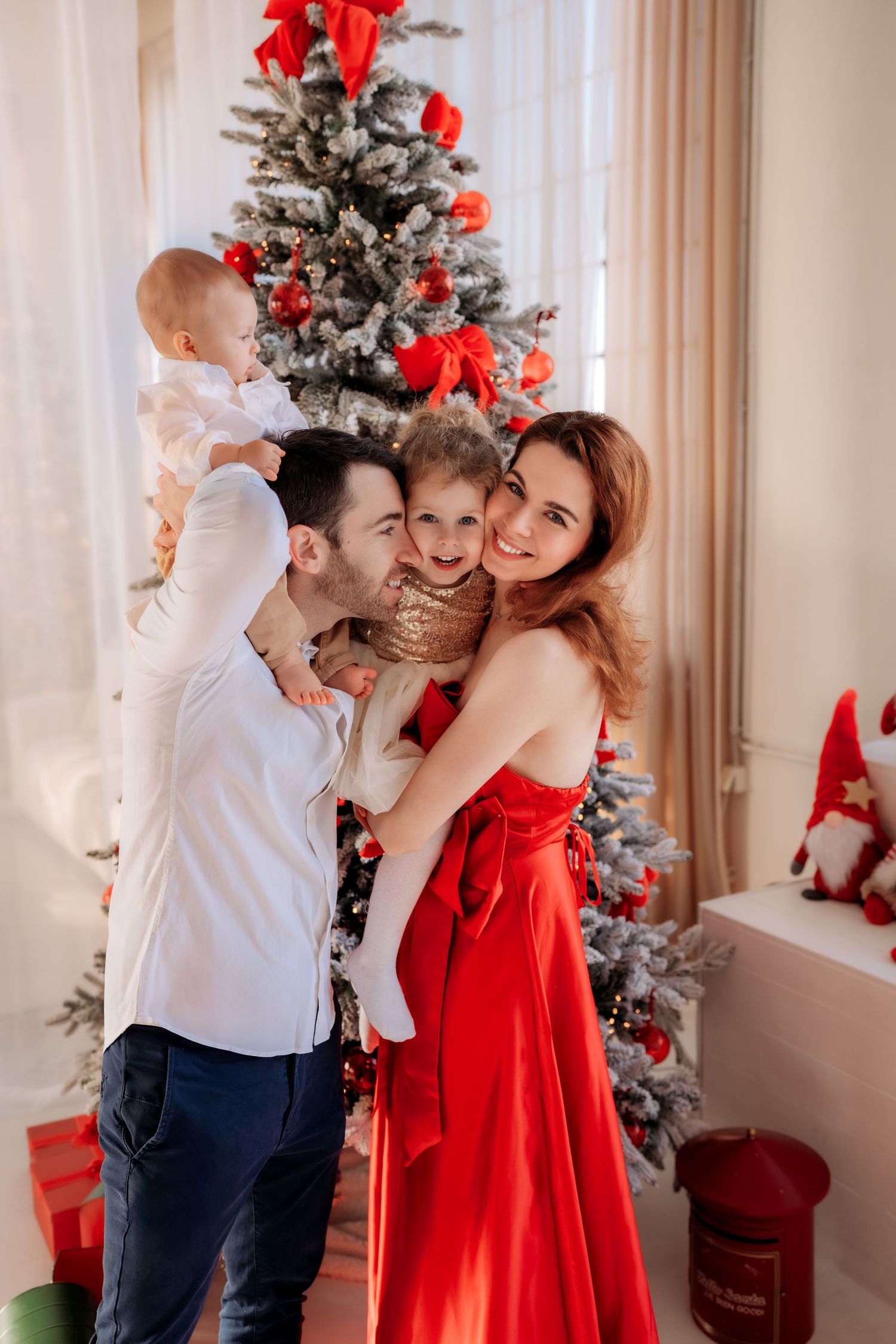 Happy family with two children posing in front of decorated Christmas tree, woman in red dress, festive holiday setting.
