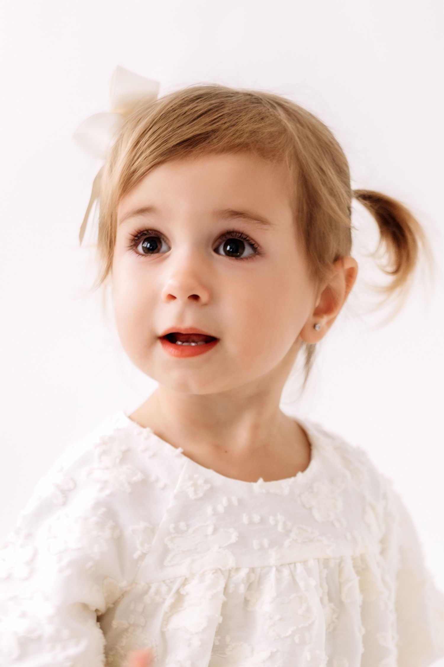 Adorable toddler girl with pigtail and white bow in white textured dress on light background.