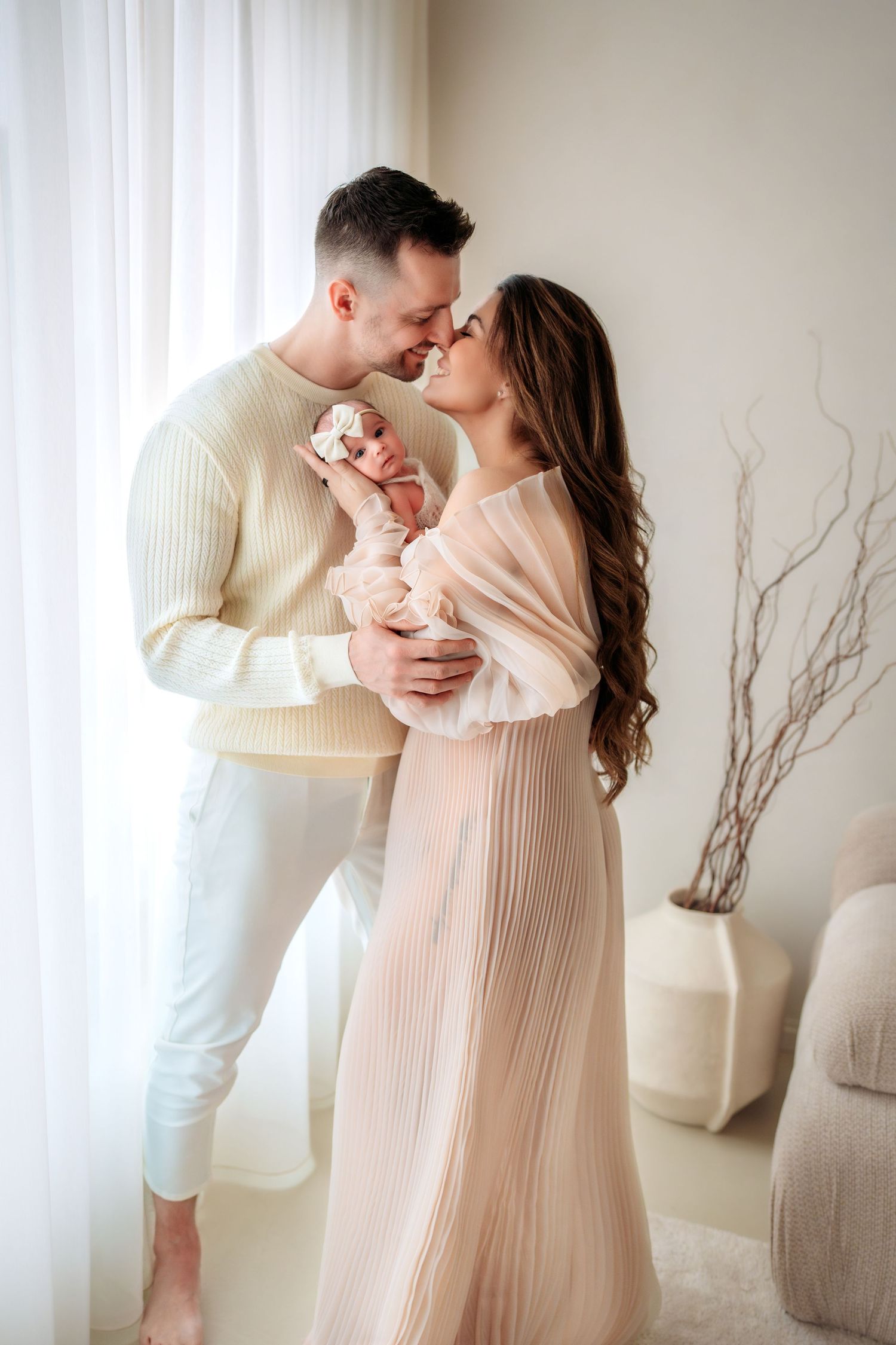 Happy couple in neutral tones sharing tender moment while holding newborn baby near bright window.