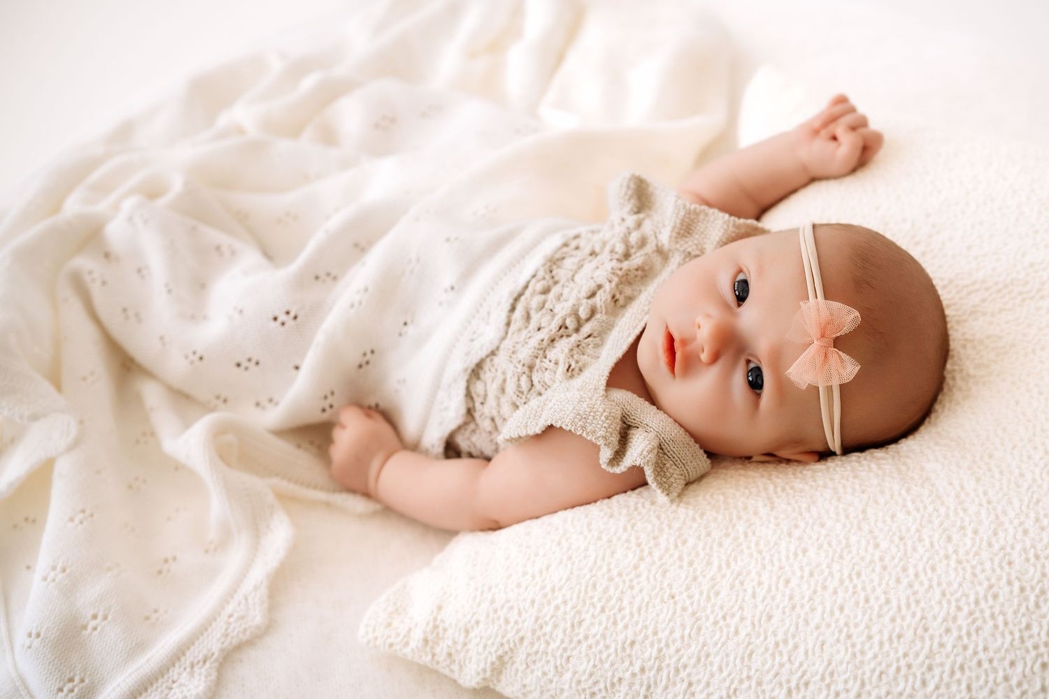 Cute baby girl in lace romper with pink bow headband wrapped in white knit blanket.