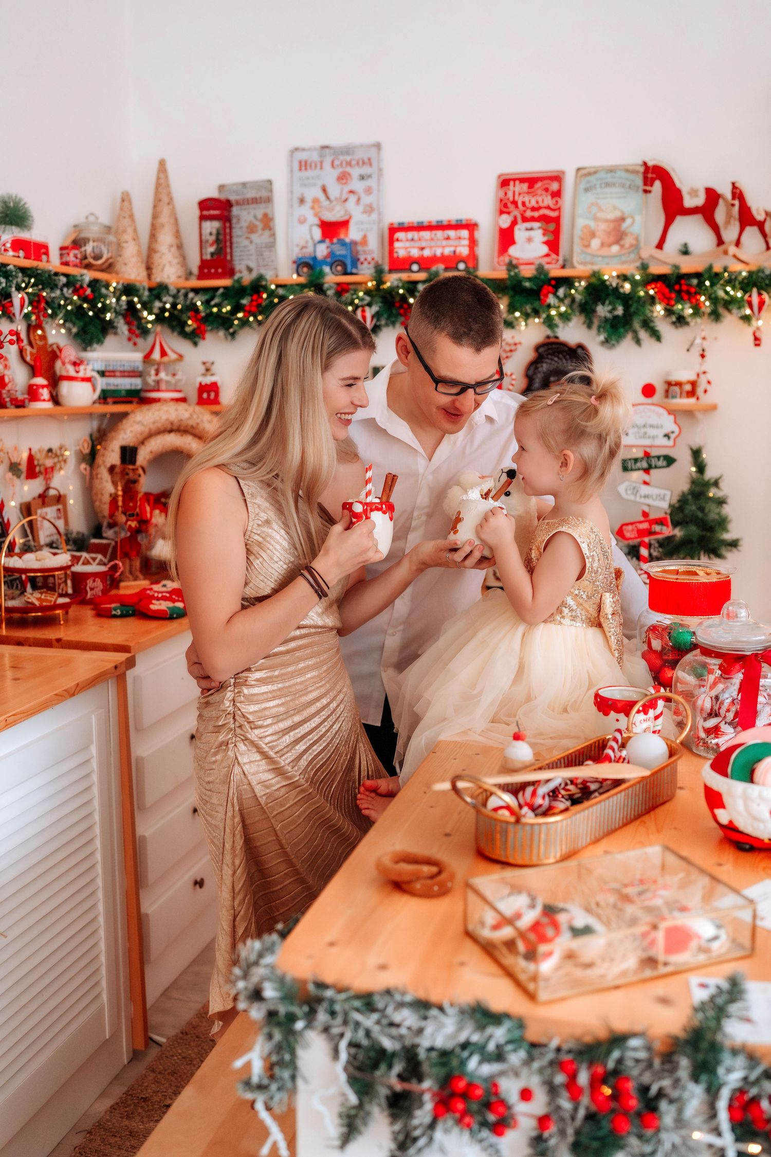Family celebrating Christmas in a festively decorated kitchen with red and gold ornaments, sharing holiday treats together.