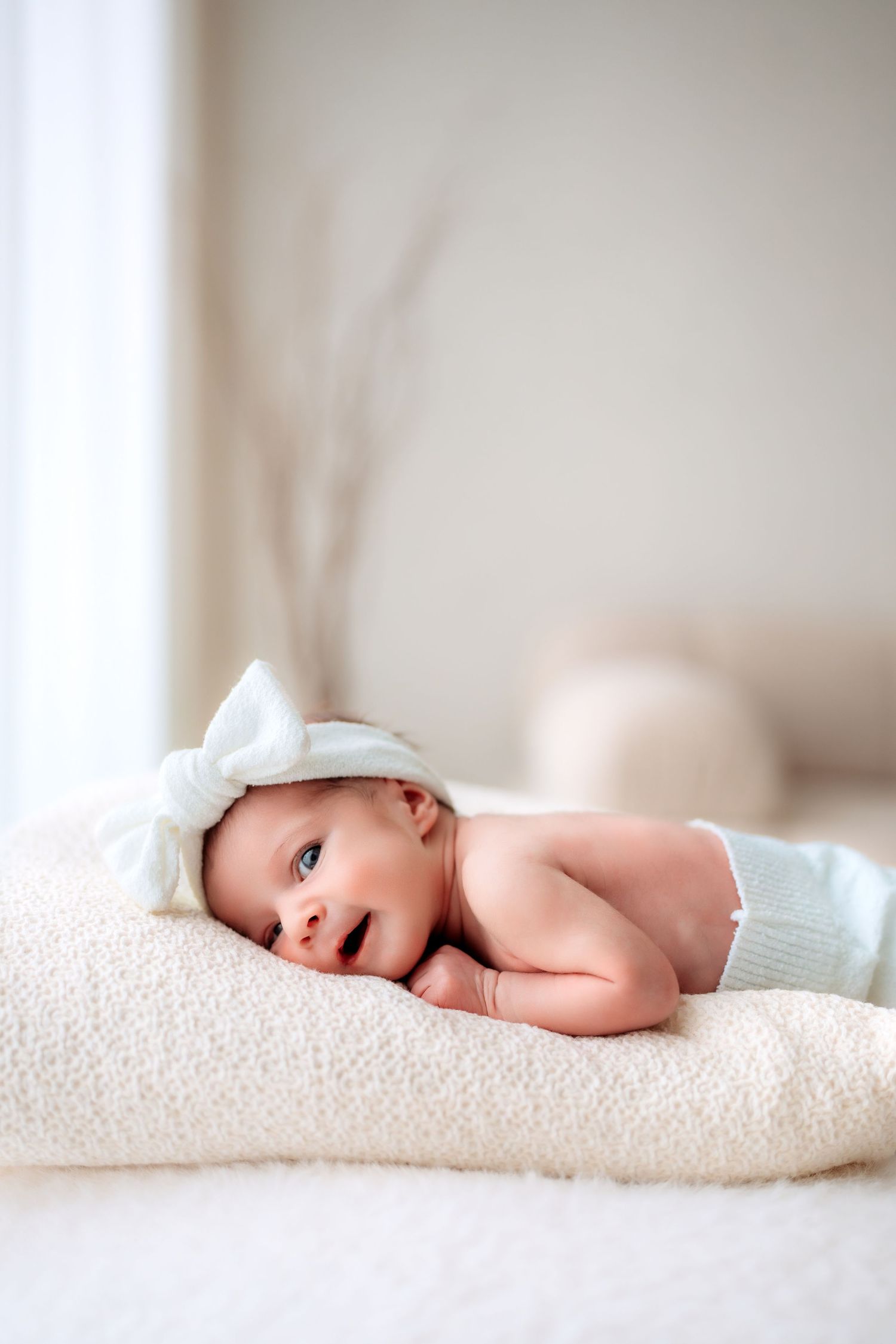 Newborn baby wearing white bow headband lying on fluffy white pillow in bright room.