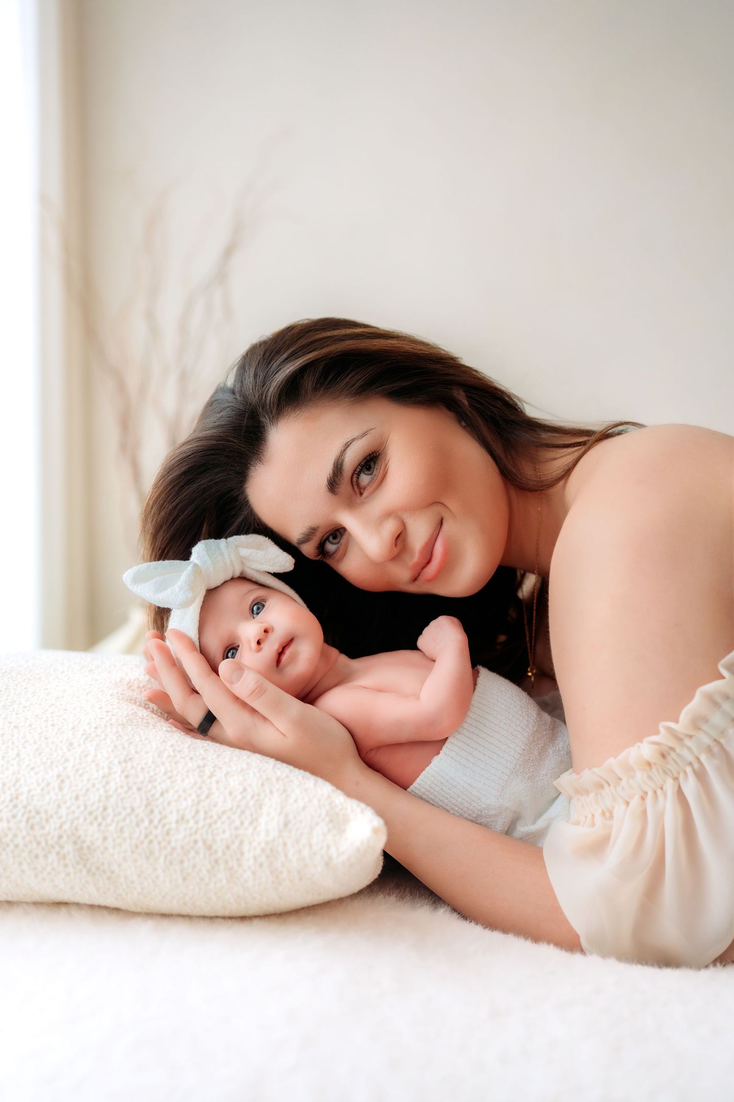 Smiling brunette mother lying beside her newborn baby with white bow on white bed.