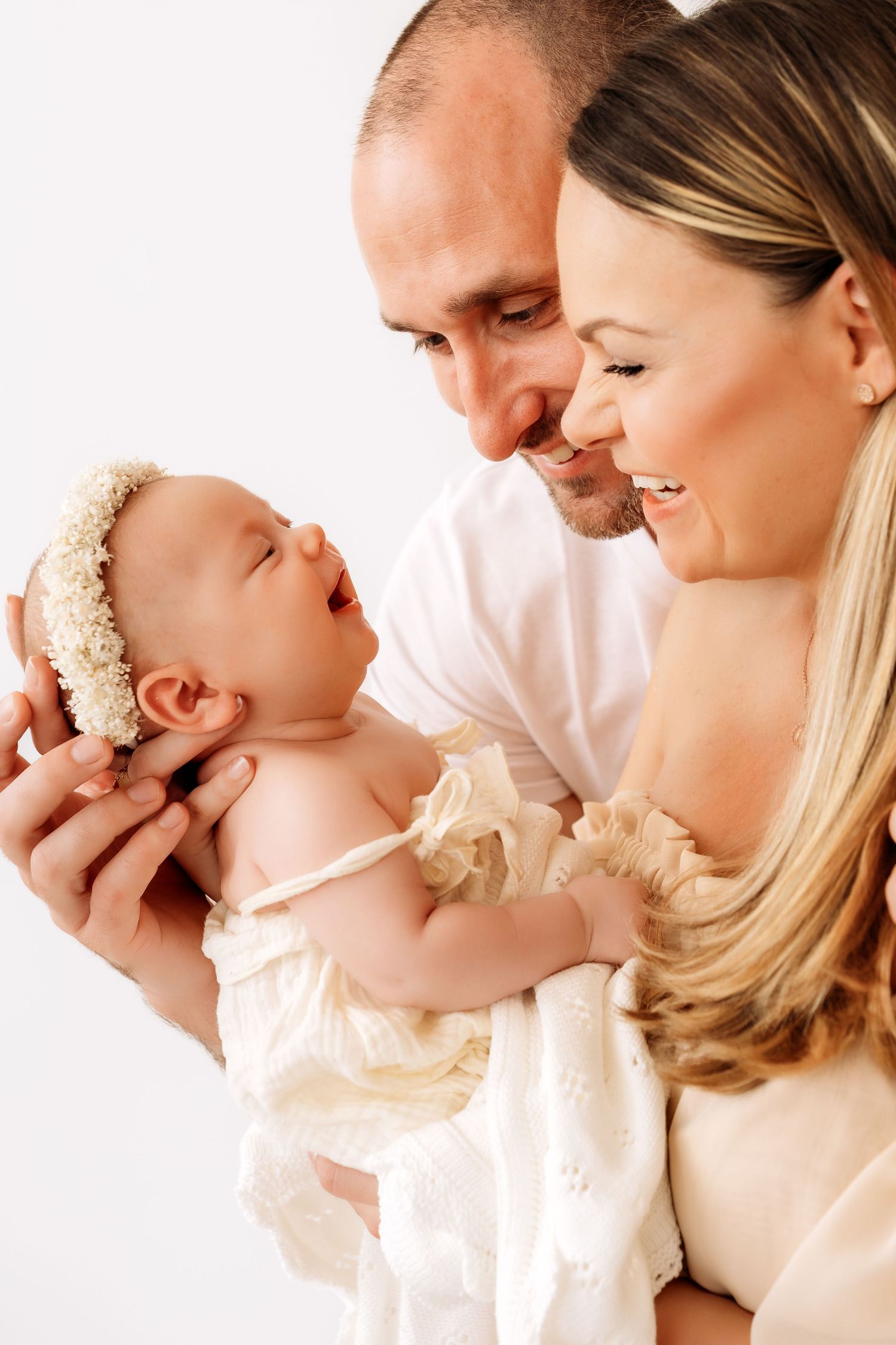 Happy parents smiling at their laughing newborn baby dressed in cream outfit.