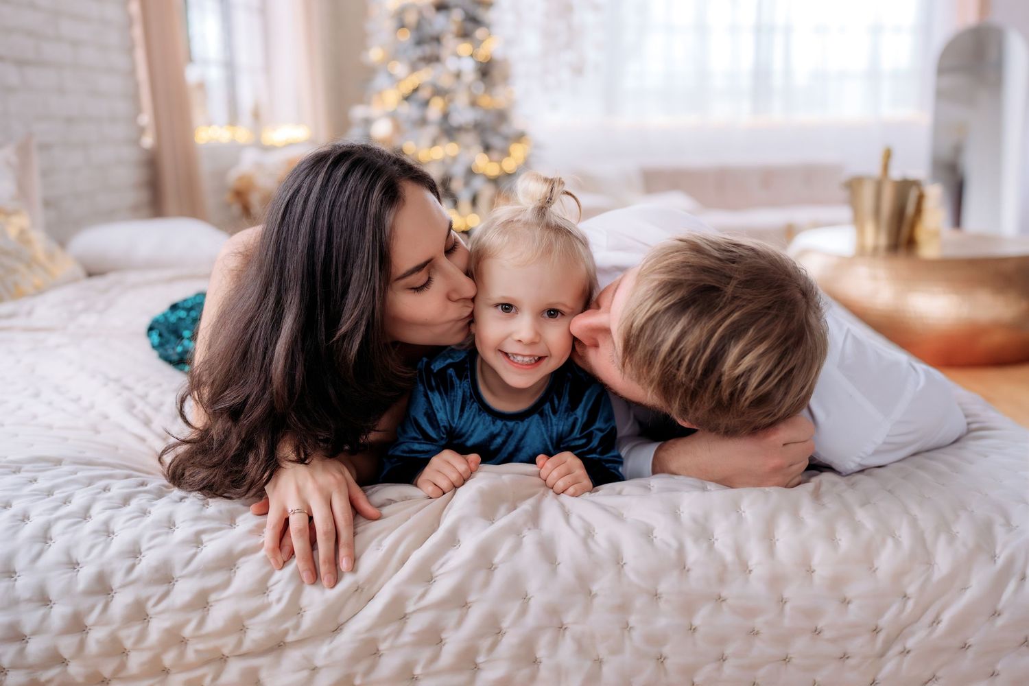Parents kissing their smiling toddler on the cheeks while lying on a bed near a Christmas tree.
