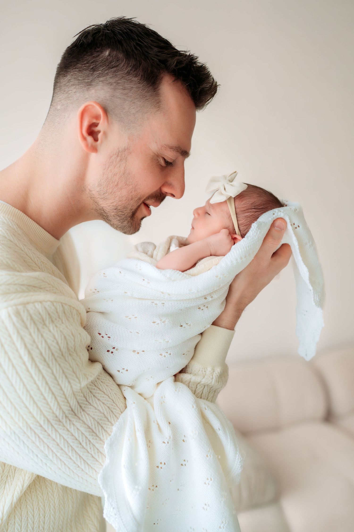 A father in a cream knit sweater tenderly holds his swaddled newborn baby close to his face.