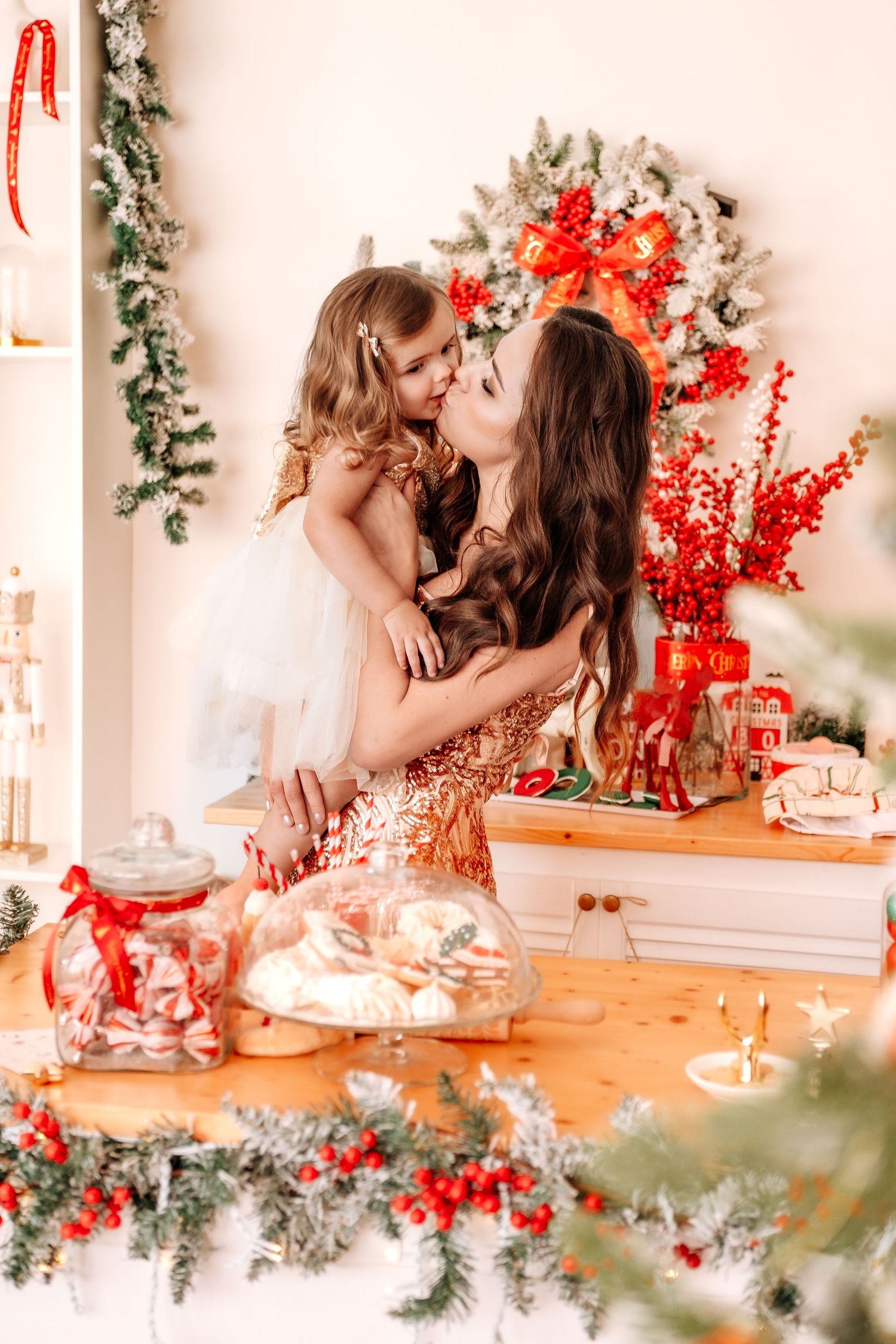 Mother and daughter sharing a kiss in a festively decorated Christmas kitchen with red and green holiday decor.