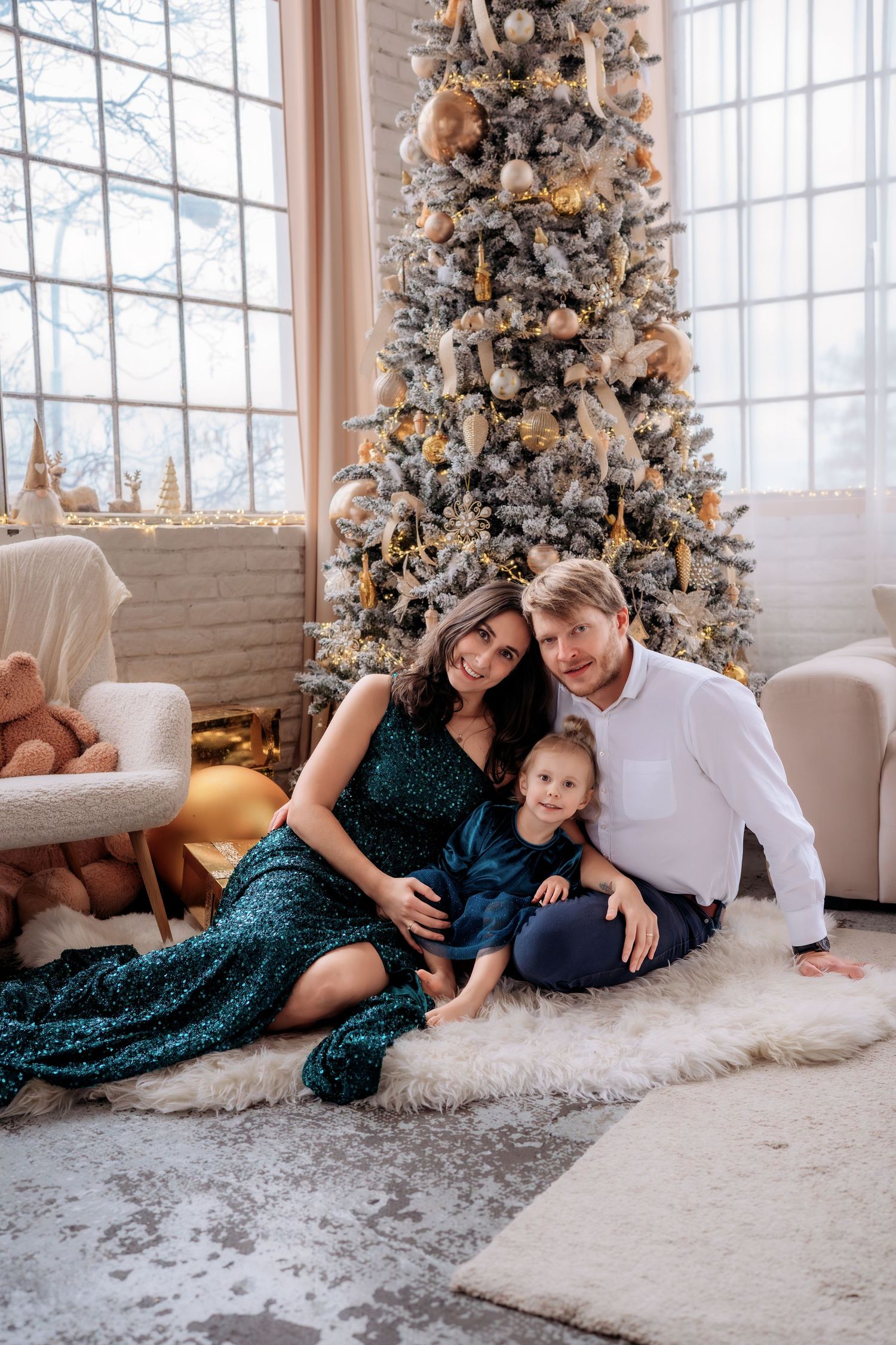 Family of three posing by decorated Christmas tree on fur rug, mother in green sequin dress, father in white shirt.