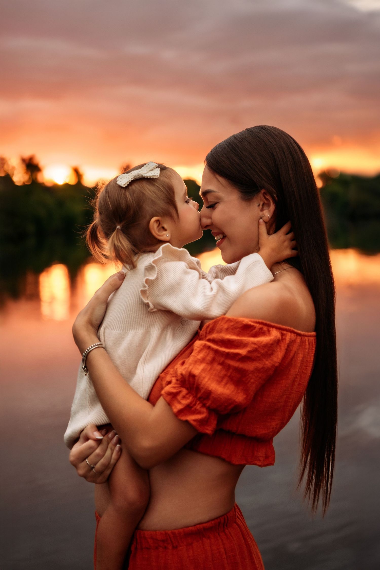 Mother and toddler daughter touching noses and laughing at golden sunset by a lake.