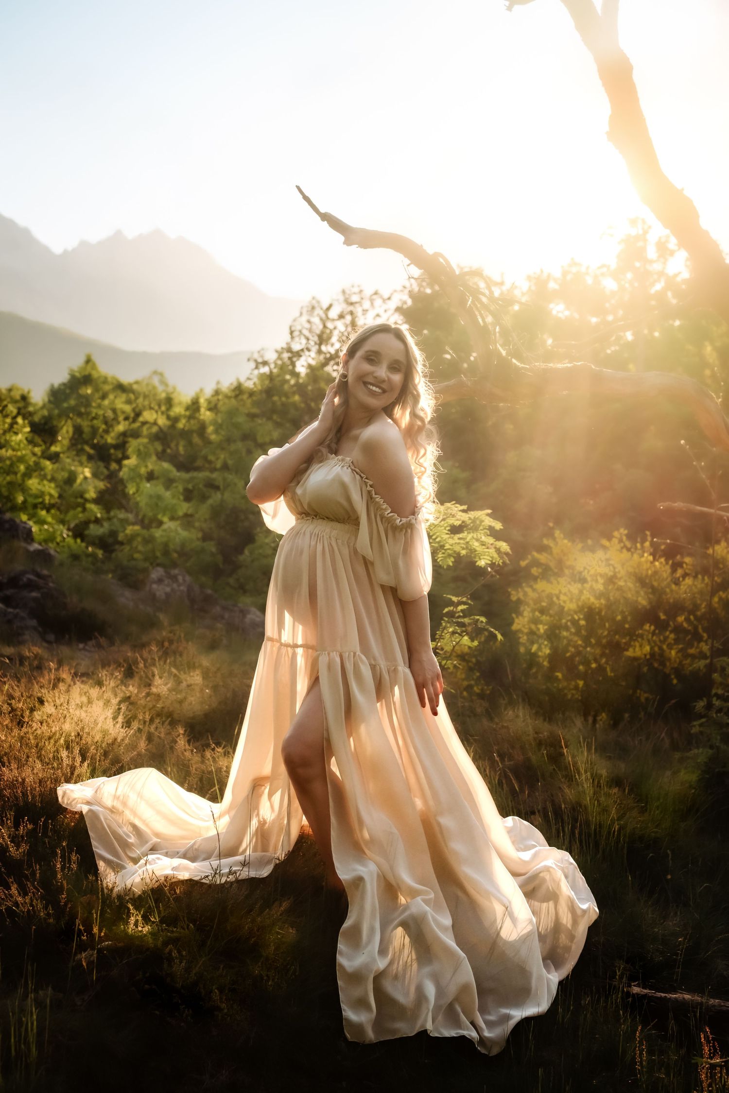 Smiling pregnant woman in flowing peach gown poses in golden sunset light among lush green trees and mountains.