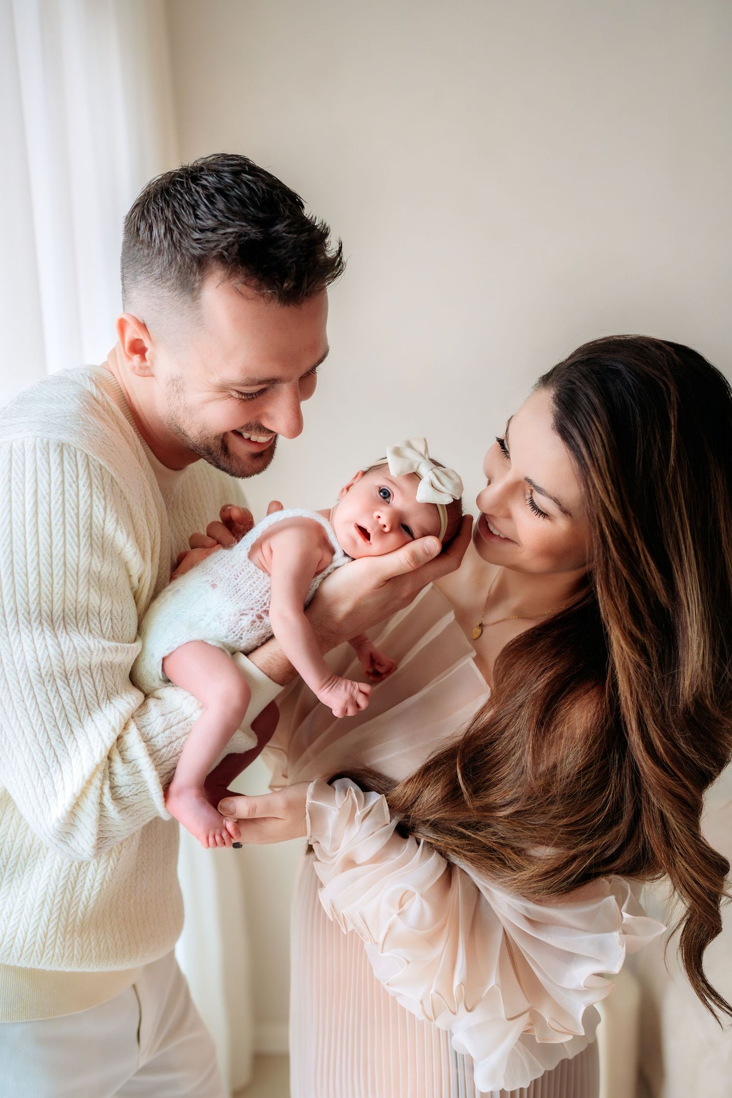 Happy parents in cream and white outfits hold and kiss their alert newborn baby girl with a bow.