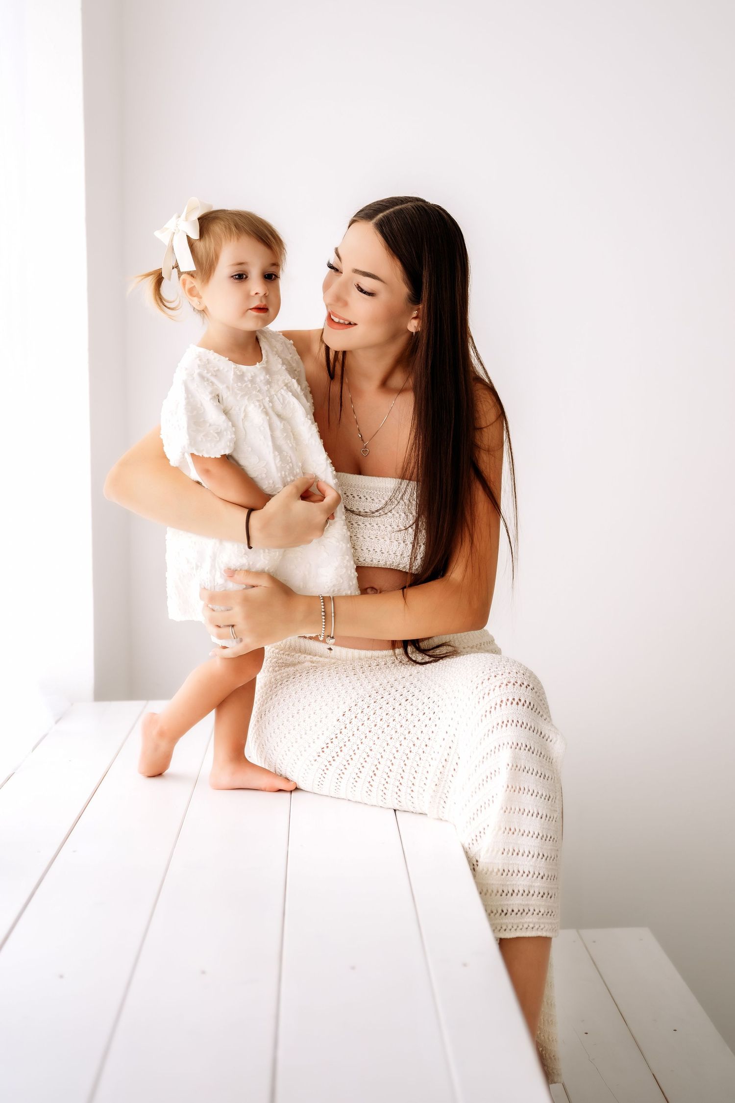 Brunette mother in white crochet outfit sitting and cuddling her toddler daughter on white bench.