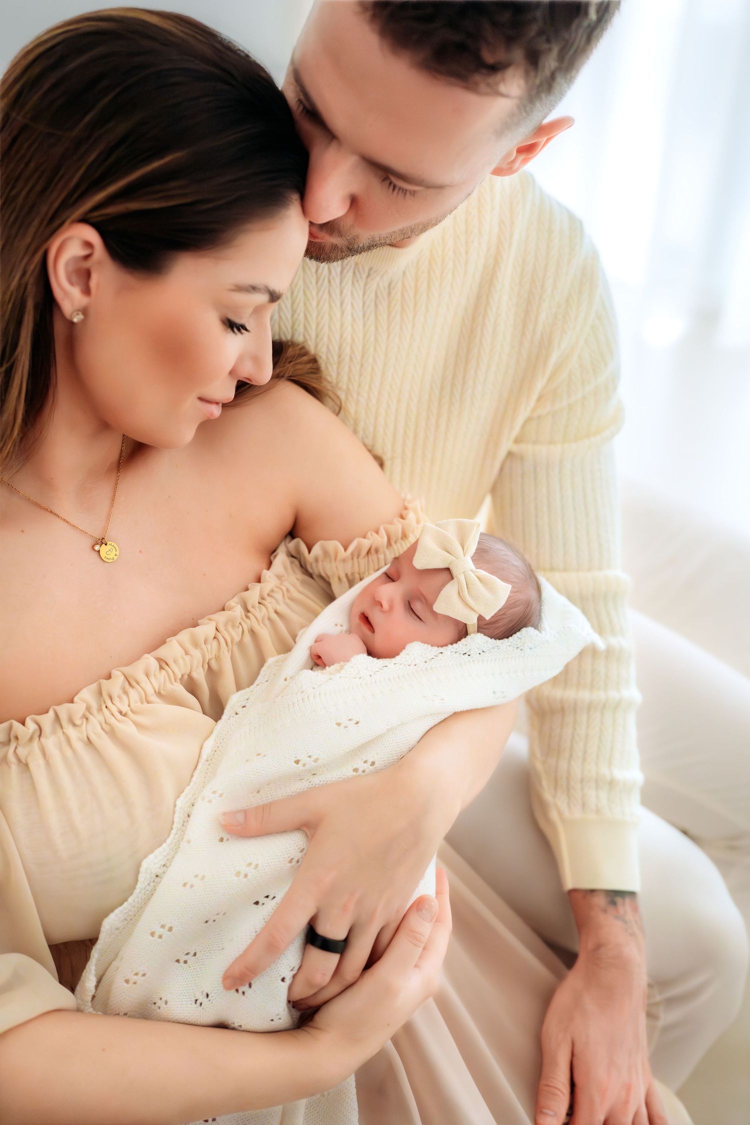 Parents in neutral tones tenderly holding swaddled newborn baby girl with white bow headband.
