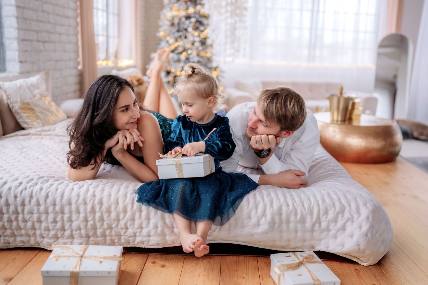 Family of three sitting on a bed opening Christmas gifts near a decorated tree in a bright festive bedroom.