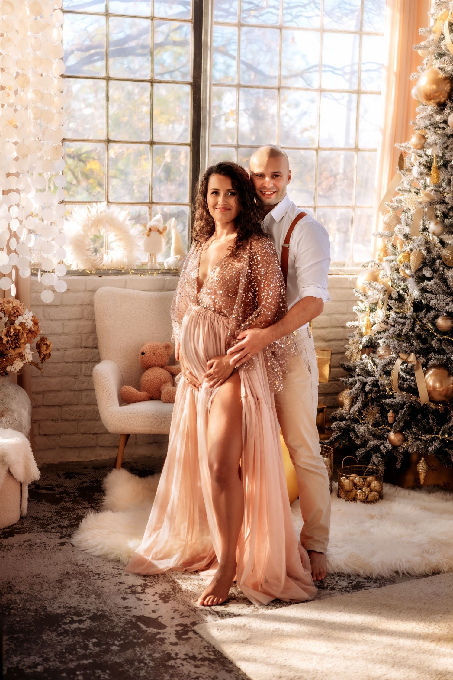 Pregnant couple posing together in a festive Christmas decorated room with a glowing tree and large window.