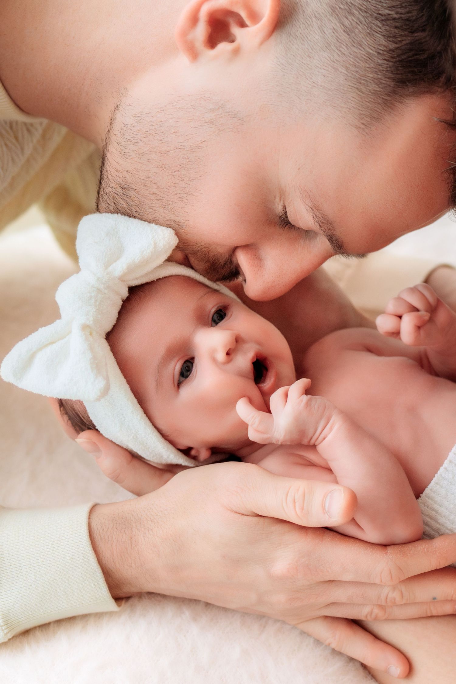 Father gently kissing his newborn baby girl wearing a white bow headband.